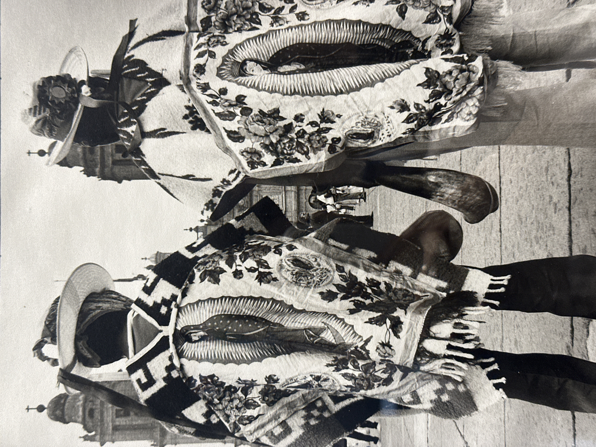 Two individuals in traditional attire stand in front of a church in Mexico City, Mexico. They wear sombreros, ponchos with an image of Our Lady of Guadalupe, and striped shawls. The building behind them is likely a historical or religious site. The scene is captured in black and white, suggesting it may be a historical photograph.