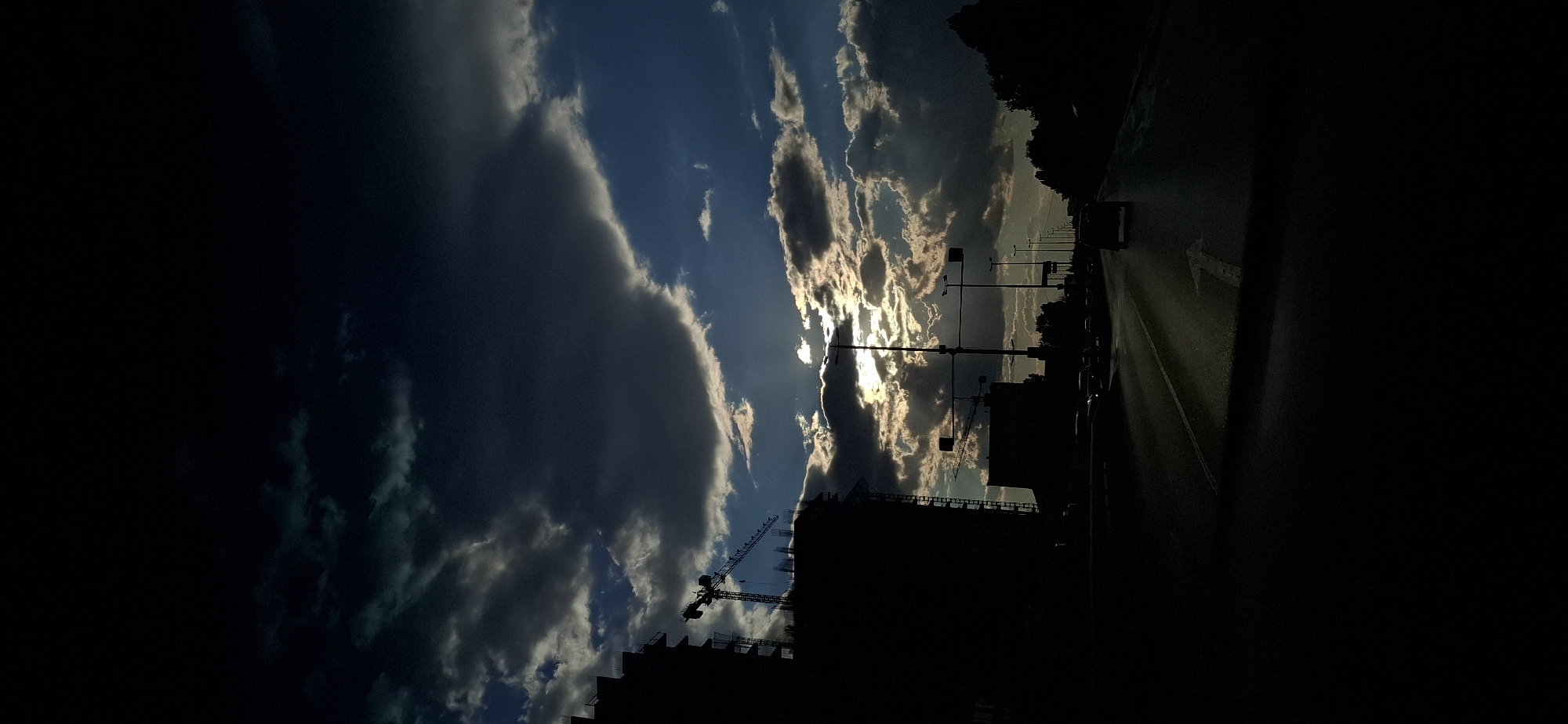 A low-angle, vertical shot captures a dusky sky with dramatic, dark clouds broken by shafts of sunlight. Below the sky, silhouetted against the bright sky, are the structures of a construction site, including a crane, and a multi-story building. Further down, a street is visible with a dark vehicle in the foreground and other vehicles in the distance, suggesting traffic. Streetlights and traffic signals are also visible along the side of the road. The scene is dimly lit, with the dark foreground contrasted by the bright sky.