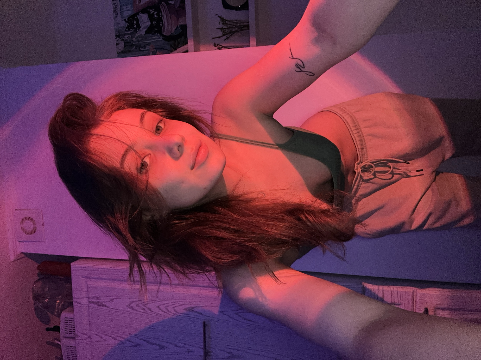 A young woman with brown hair pulled up in a bun is lying on a bed. She is wearing a green top and tan sweatpants. She has a tattoo on her left arm. The room is lit with pink and purple light. There is a dresser and light switch visible in the background, and there are framed pictures on a wall. The location is not specified in the image and there is no visual information to ascertain that the location is in Moscow, Russia.