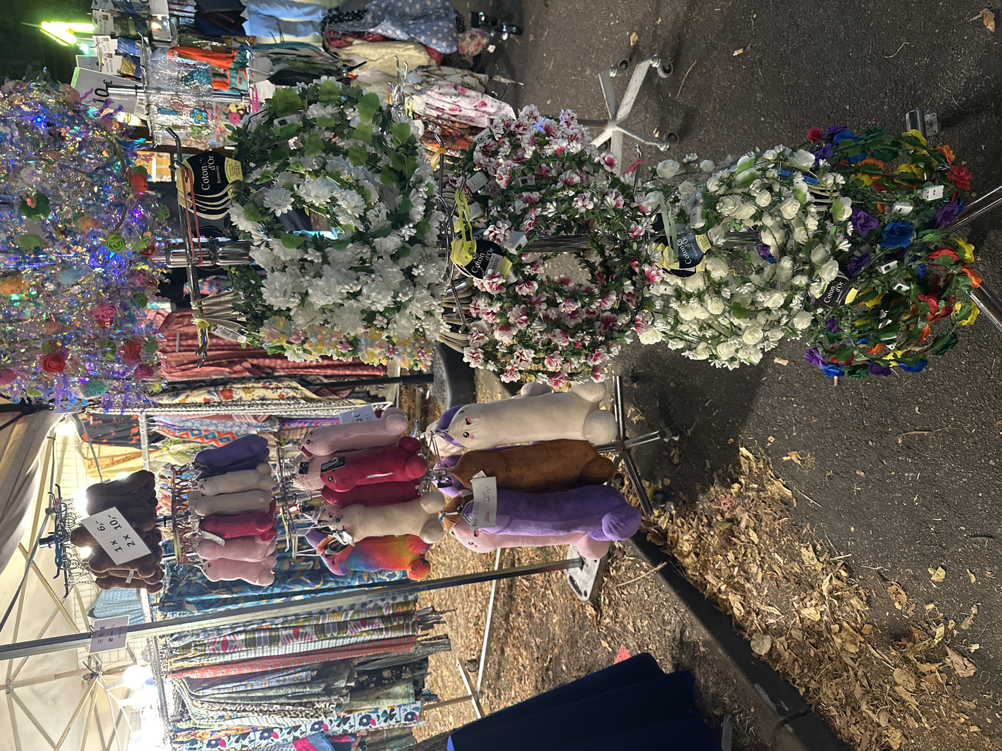 The image shows an outdoor market stall in Bruchhausen-Vilsen, Germany, displaying various items for sale. In the foreground, there are several plush phallic-shaped toys hanging from a metal rack. Above these toys, on the same rack, are two circular floral wreaths, one predominantly white with green leaves and another with pink and white flowers. Further up the rack, there are more floral wreaths adorned with colorful small flowers. The background reveals racks of clothing, some with patterns and vibrant colors, and a sparkling, multi-colored decorative canopy hanging overhead, likely part of the stall's lighting or decoration. Fallen leaves are visible on the ground in front of the stall. The lighting suggests it is nighttime or late evening.