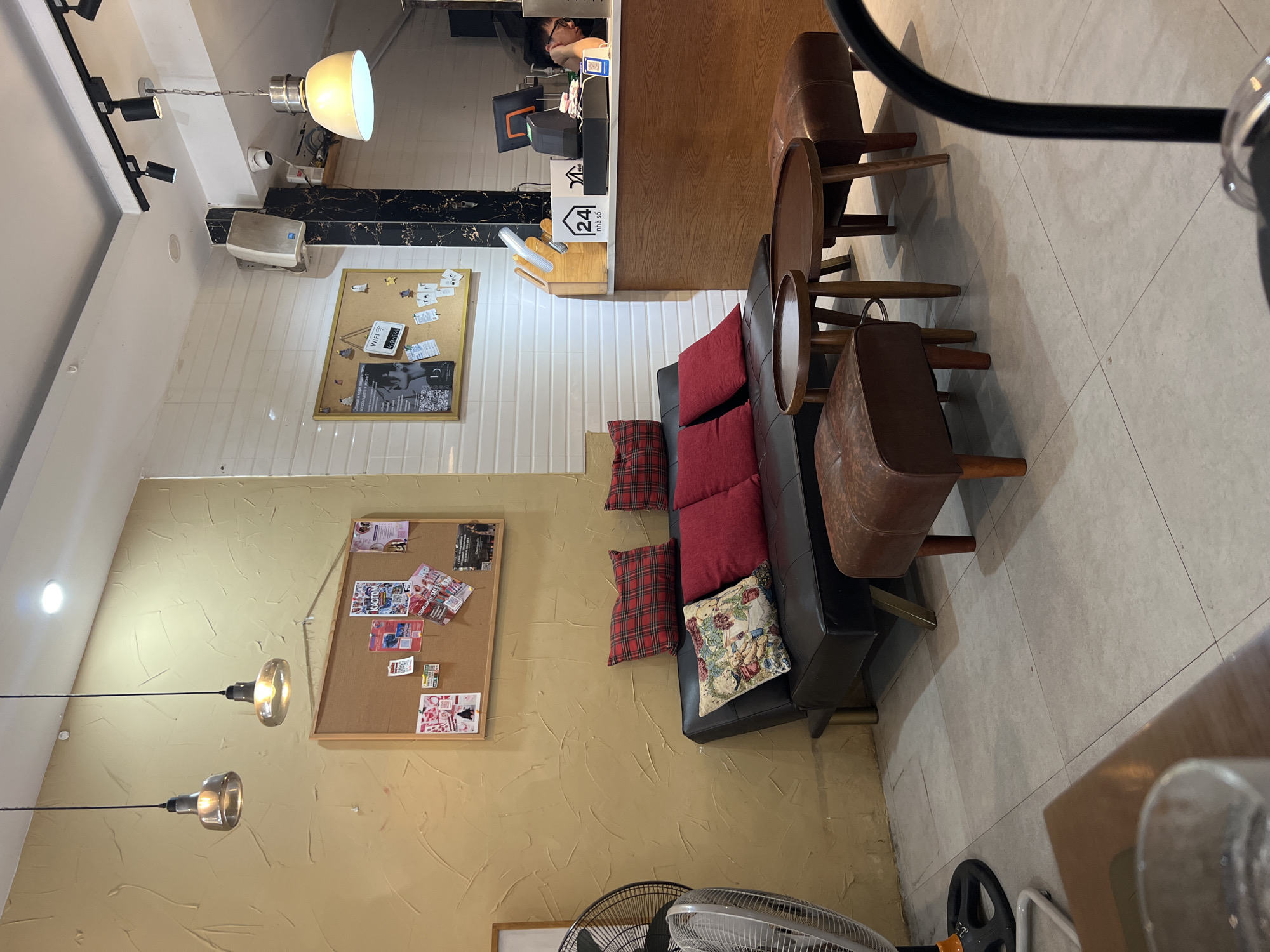 The image depicts the interior of a cafe or lounge area. In the foreground, to the left, is a portion of a glass on a wooden table. The main subject of the image is a black leather sofa with red plaid pillows and one decorative pillow. In front of the sofa are two small wooden tables with two brown leather ottomans. To the left of the sofa, a large silver floor fan is visible. The wall behind the sofa is painted a textured mustard yellow and features a corkboard with various papers pinned to it. To the right of the yellow wall is a white tiled wall with another corkboard. A wooden counter with a cash register and a person working behind it is visible in the background. The ceiling has track lighting and pendant lights. The floor is tiled with light gray tiles. The overall impression is a cozy, casual seating area within a commercial establishment.