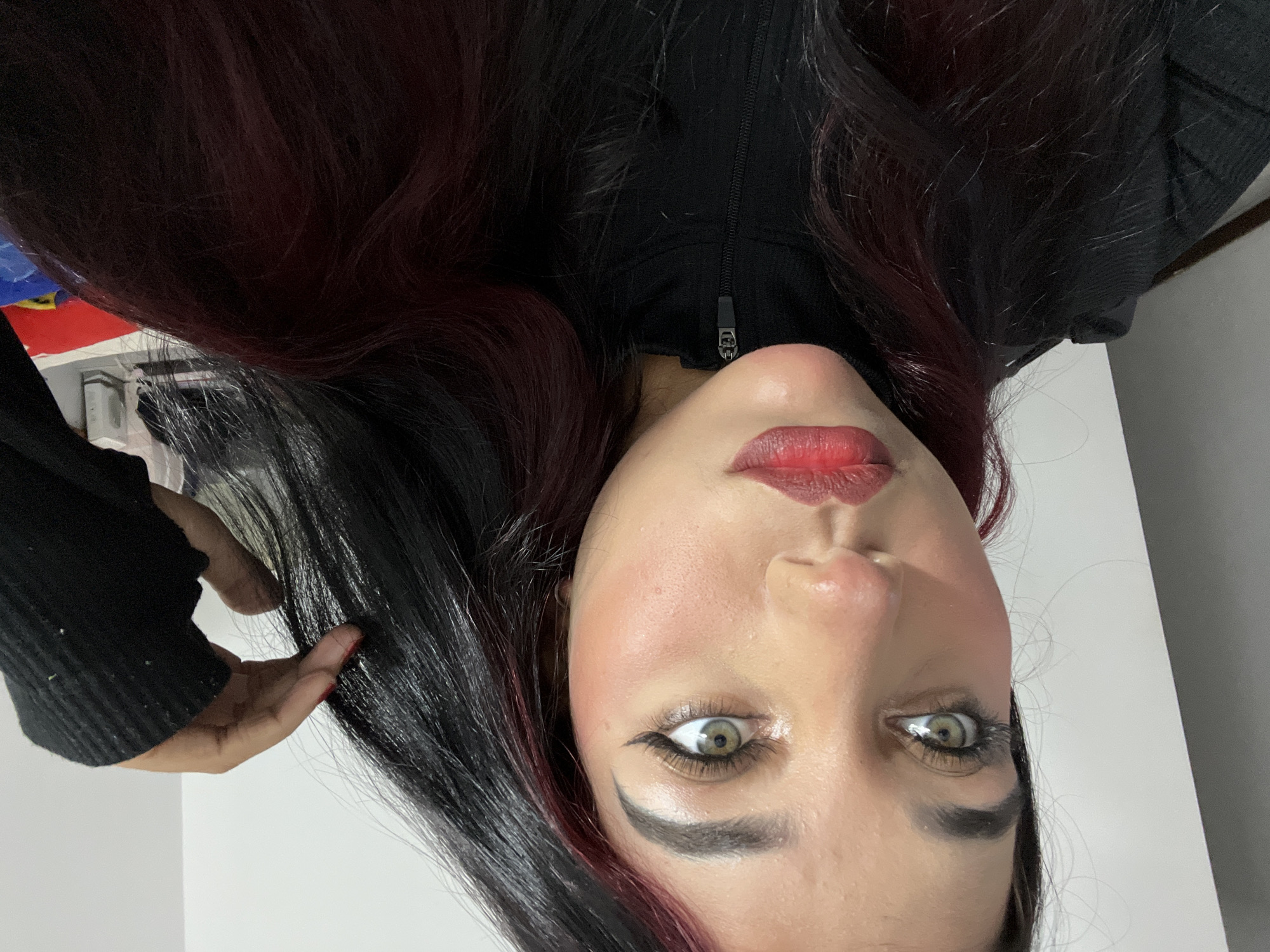 A medium shot shows a young woman looking to her left with her hand touching her hair. She has light brown hair with streaks of red, dark eyebrows, and is wearing makeup including eyeliner and lipstick. She is wearing a black turtleneck. The background is a plain white wall and part of a gray wall. The image appears to be taken indoors. There is no visible indication of the image being taken in Porlamar, Venezuela.