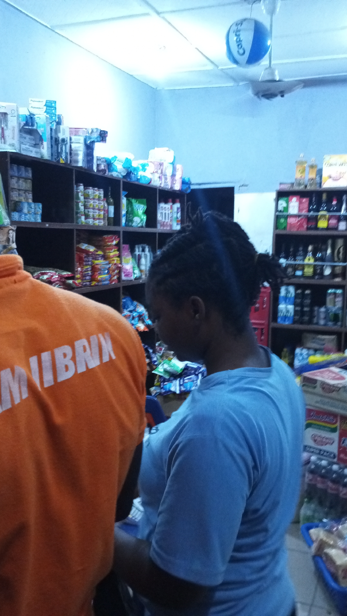 The photograph depicts the interior of a small retail store in Adankolo, Nigeria. Two individuals are present. The foreground is dominated by a person wearing an orange shirt with the word "MUIBRNK" printed on the back in white letters. Standing beside the person is another individual, facing away from the camera and wearing a light blue t-shirt. This person has dark hair styled in braids and a ponytail.
Behind the individuals are shelves stocked with various items. The shelving units contain a variety of consumer products, including canned goods, snack foods, and bottled beverages. A blue and white beach ball hangs from the ceiling near a white ceiling fan. The store's walls are pale blue.