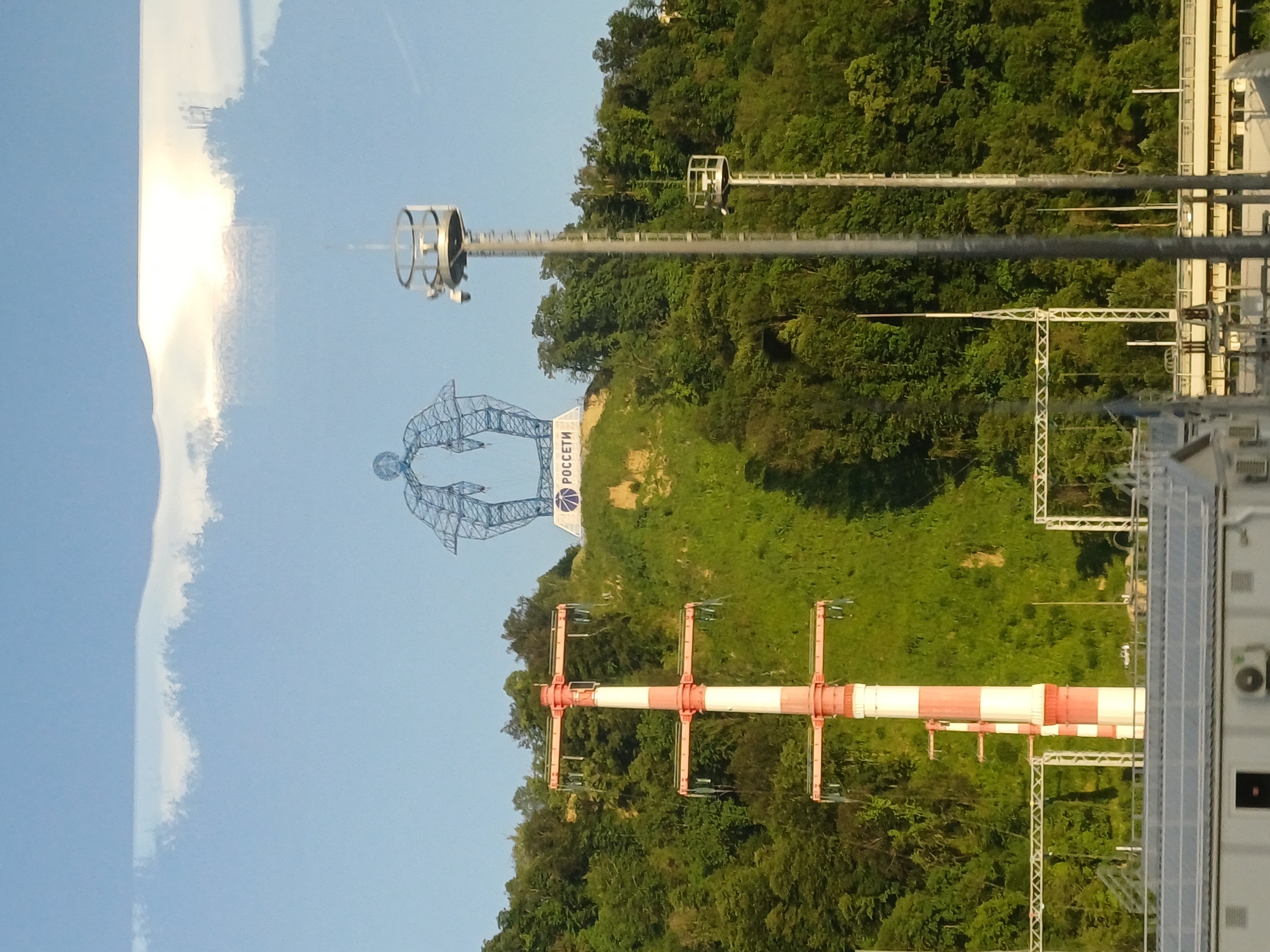 In the city of Vysokoye, Russia, a metal sculpture of two figures embracing is situated atop a grassy hill. The sculpture features a logo that reads "РОСЕТИ". In the foreground, a tall, red and white striped electrical pylon stands prominently. Two surveillance towers with circular platforms and cameras are also visible on the right side of the image. The environment is characterized by a clear blue sky with a thin layer of clouds, and the hill is covered with dense green foliage. In the lower part of the image, parts of industrial buildings and infrastructure can be seen.