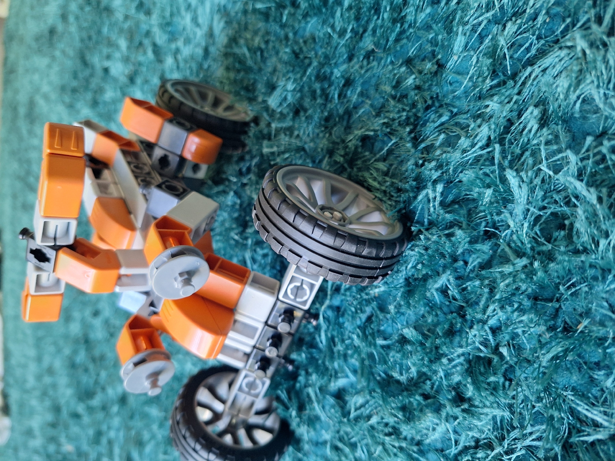 An orange and gray Lego ATV is positioned on a teal, shag carpet. The ATV has four wheels with silver rims and black tires. The front of the ATV has a light gray bar with two circular gray pieces attached, and orange curved pieces extending outwards. The main body of the ATV is constructed with orange and gray Lego bricks, forming a boxy structure. The rear of the ATV is also made of Lego bricks, with orange pieces at the top and gray pieces forming the chassis. The vehicle is viewed from a low angle, with the right front wheel closer to the viewer. The background is entirely composed of the teal carpet, with some areas slightly out of focus. There is no indication of Varna, Bulgaria, or any specific city location.