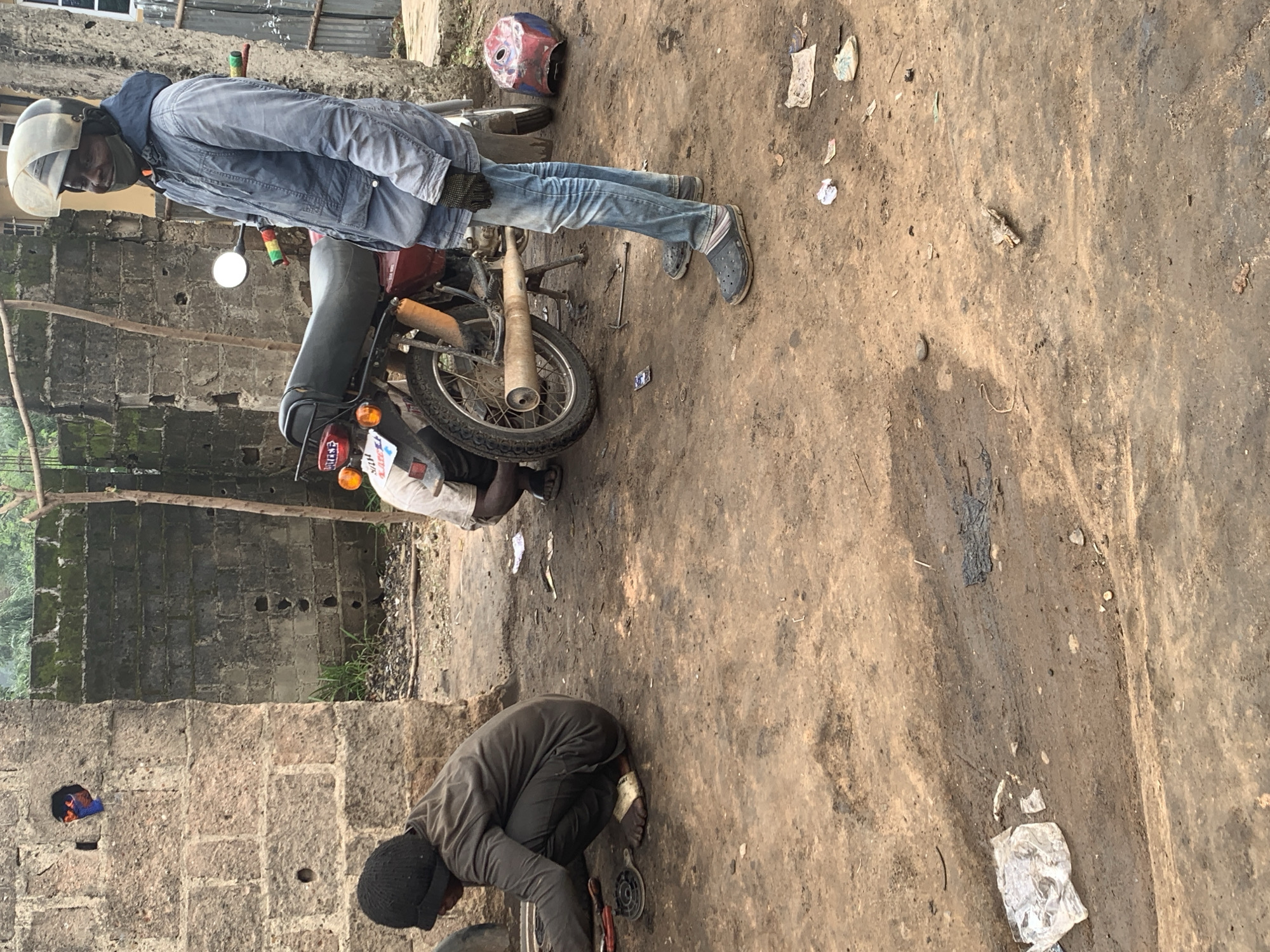 The image depicts an outdoor motorcycle repair scene in Shagamu, Nigeria. Two individuals are visible; one, wearing a black beanie and dark clothing, is crouched down and appears to be working on a motorcycle part. The second individual, standing and wearing a blue jacket, jeans, and a helmet, is looking towards the camera and smiling. A red motorcycle with a brown seat is the central object of the scene, with its exhaust pipe visible. The environment is dusty and appears to be a workshop or informal repair area, with a brick wall and a rudimentary shelter structure in the background. Scattered debris, including plastic bags and tools, are present on the ground, indicating ongoing activity.