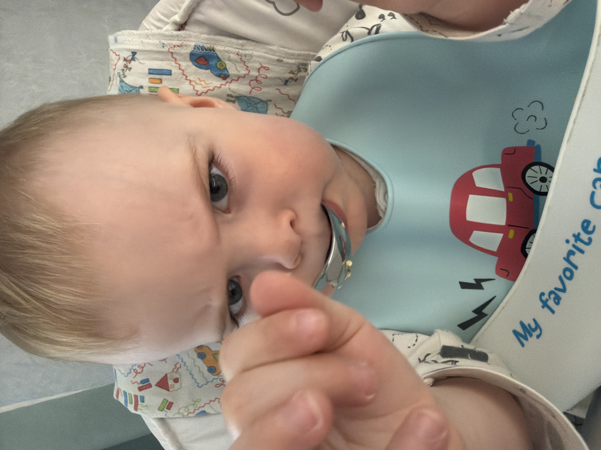 A baby with light blonde hair, gray eyes, and fair skin is the primary subject of the image. The baby is wearing a light blue bib with a red car design on it, along with a patterned fabric around the neck area. A metal spoon is partially visible in the baby's mouth. The background appears to be a light-colored interior. The scene is implied to be set in Votkinsk, Russia.