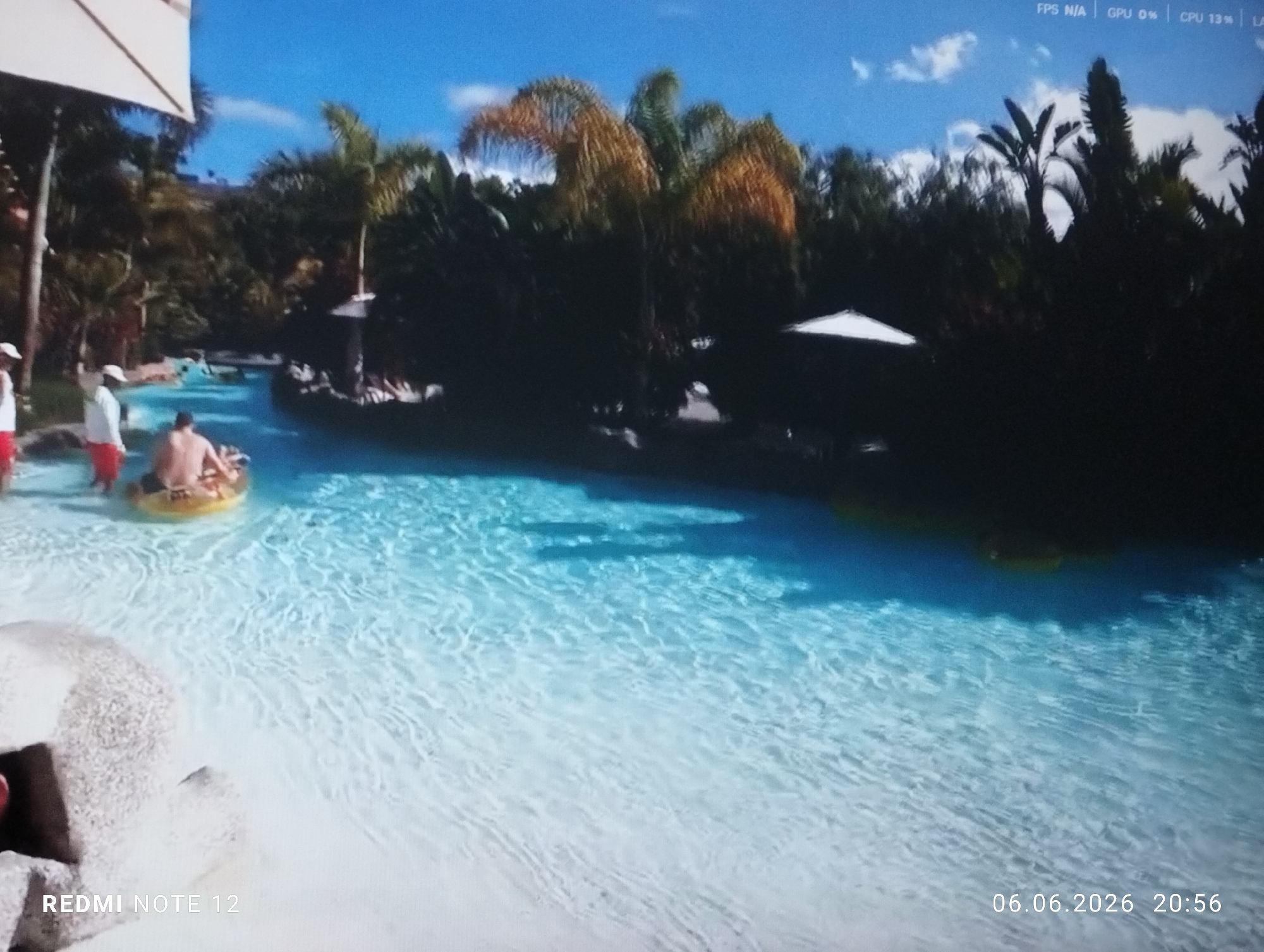 The image depicts a photograph of a screen displaying an outdoor recreational scene, likely a water park or resort.
**Environment:** The scene features a large, light blue body of water, possibly a lazy river or a shallow pool, which occupies the lower half of the frame. The water exhibits rippled surface textures, indicative of movement or shallow depth. The edges of the water appear to be a light, sandy color. Beyond the water, a dense tropical or subtropical landscape is visible, dominated by numerous palm trees and thick green foliage, creating a lush backdrop. A bright blue sky with scattered white clouds is visible in the upper portion of the display.
**Subjects and Objects:**
* **People:** On the left side of the water, near the edge, several individuals are present. One person, positioned centrally in the water, is seated in a yellow inflatable ring, facing away from the camera. To their left, two individuals wearing red shirts and white caps, possibly lifeguards or staff, stand at the water's edge. Other indistinct figures are visible further in the background water channel.
* **Structures:** A large, light-colored umbrella or canopy structure is partially visible in the upper-left quadrant, providing shade over an unseen area. Further back in the water channel, a dark, boat-like structure, possibly a themed decoration or a ride element, is present. A smaller, conical white structure is dimly visible amidst the foliage in the background.
* **On-screen Display Elements:** Various digital overlays are present on the screen being photographed:
* "REDMI NOTE 12" is visible in the lower-left corner.
* "06.06.2026 20:56" is displayed in the lower-right corner, indicating the date and time the photograph of the screen was captured.
* In the upper-right corner, performance metrics are visible: "FPS N/A | GPU 0% | CPU 13% |".
**Actions and Interactions:** The individuals at the water's edge appear to be standing or observing. The person in the inflatable ring is passively floating in the water, suggesting leisure or recreation.
**Location:** There are no discernible landmarks, signs, or architectural features within the displayed image content to definitively identify the specific location as being within the city of Burgas, Bulgaria. The environment presented is typical of many tropical or subtropical water parks globally.