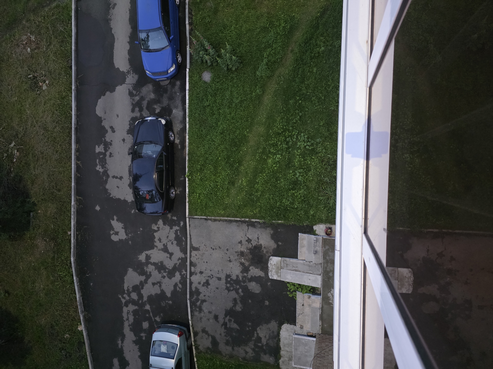 An aerial view captures a residential courtyard in Chelyabinsk, Russia. Three parked cars are visible: a silver sedan on the left, a black sedan in the center, and a blue station wagon on the right. The cars are positioned on a wet asphalt surface adjacent to a strip of green grass. A concrete pathway with steps leads from a building's foundation towards the grass. The foreground is dominated by the reflective surface of a window or balcony enclosure.