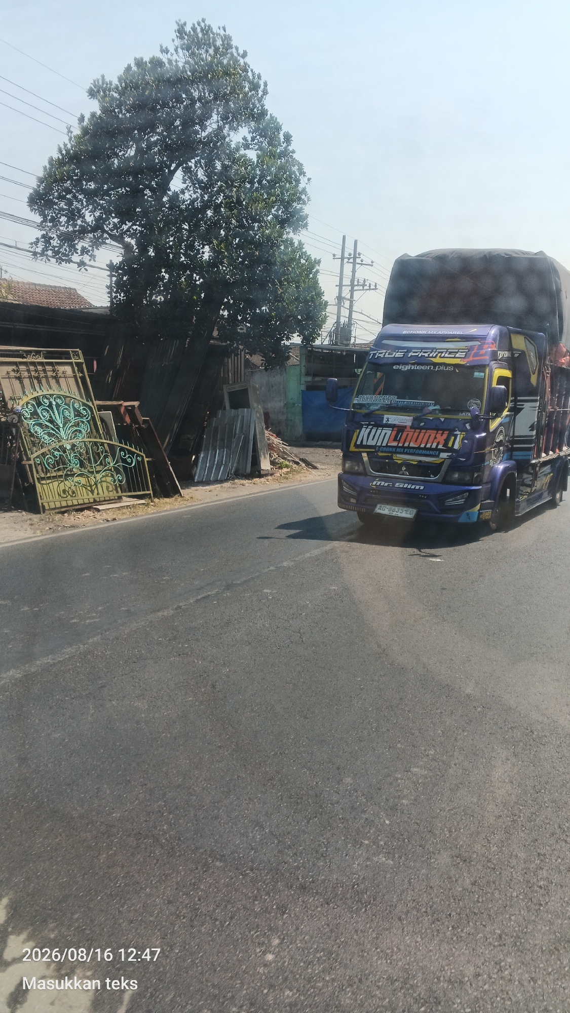 A blue and yellow truck is driving down a paved road in Pakis, Indonesia. The truck has various decals on its side, including "TRUE PRINCE" and "KUN LAUNX". It is carrying a large, dark-colored load on its back, covered with a tarp. To the left of the road, there are several metal gates leaning against a building, along with some corrugated metal sheets and other discarded items. A large tree with dark green foliage stands behind the gates and building. Power lines are visible in the background against a clear sky. The timestamp "2026/08/16 12:47" and the text "Masukkan teks" are visible in the bottom left corner of the image.