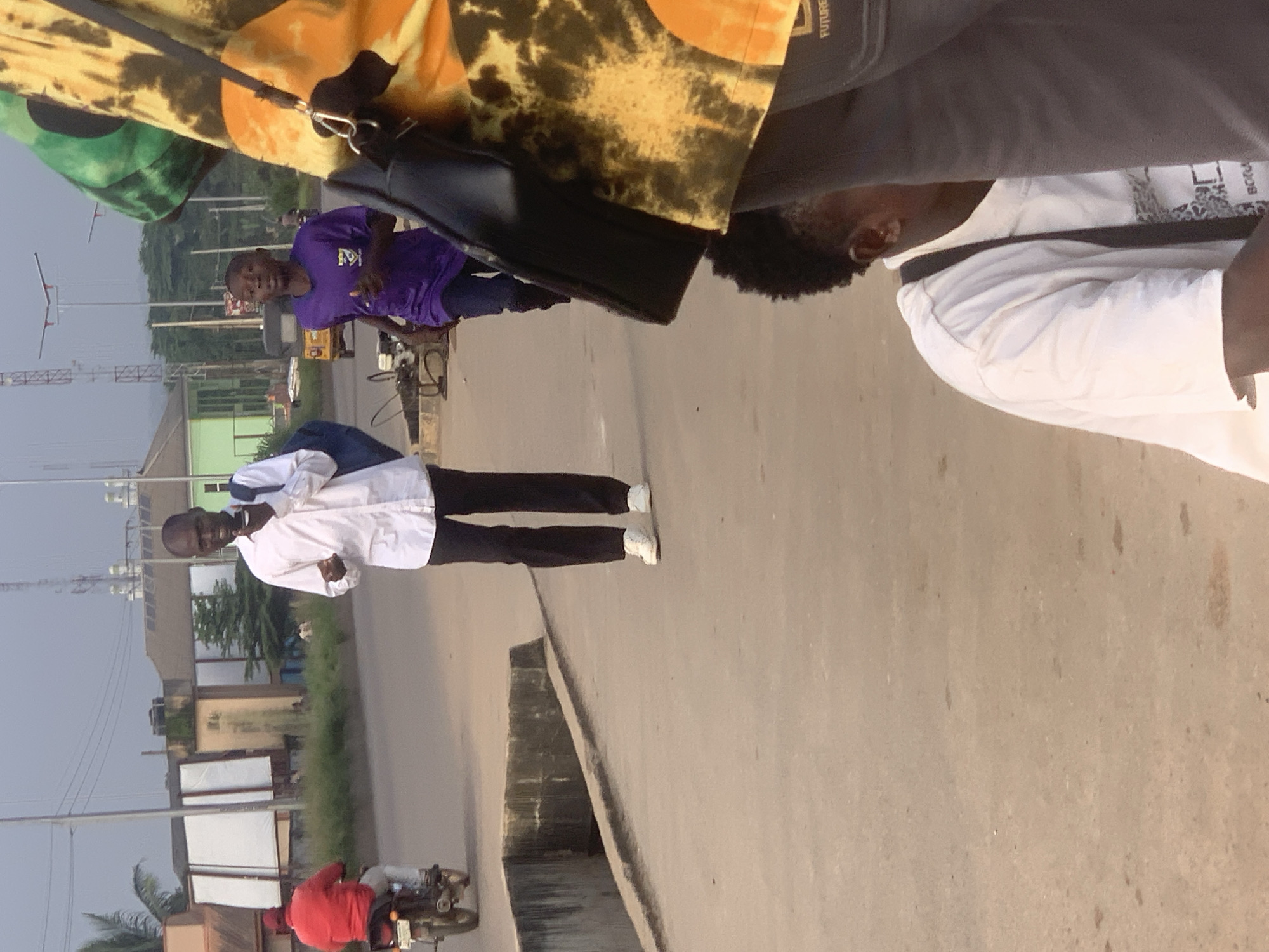 The image depicts a street scene in Shagamu, Nigeria, under an overcast sky. In the foreground, a person wearing a white t-shirt and dark pants walks on a paved walkway, with a dark bag slung over their shoulder. To their right, partially visible, is another person wearing a colorful tie-dye garment and carrying a black bag. Further into the scene, a young person in a purple t-shirt runs on the road. Behind them, a person in a red shirt rides a motorcycle. Buildings line the side of the road, with some appearing to be under construction or renovation, and a visible power line pole with multiple cables. A small yellow vehicle, possibly a tuk-tuk, is also present on the road. The walkway is elevated and appears to be next to a drainage ditch.