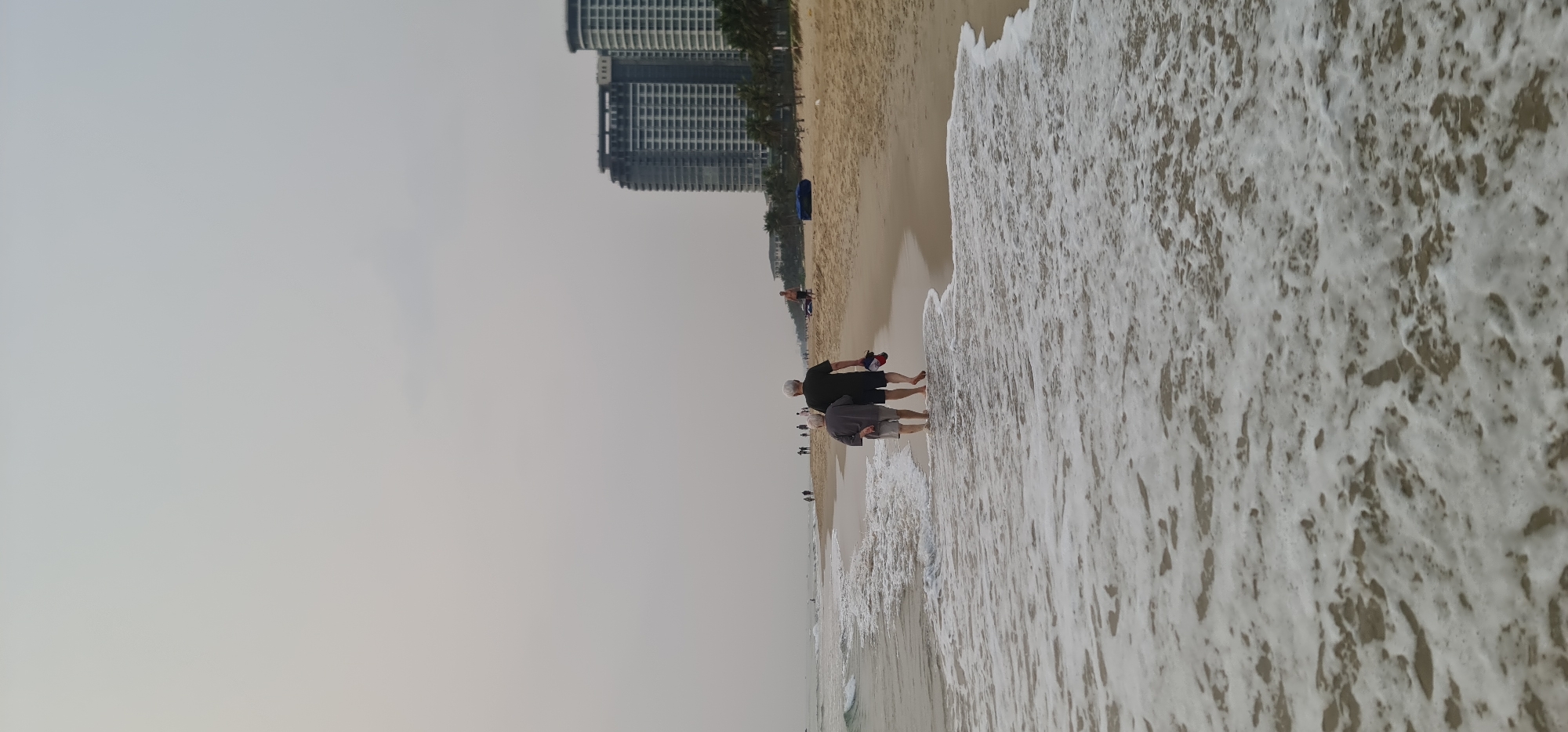 The scene captures a sandy beach in Đà Nẵng, Vietnam. The shore curves slightly, meeting the ocean's edge where foamy waves churn. Two modern, high-rise buildings stand in the background. Several people are walking along the beach, with two men in the foreground. The sky is overcast and grey.