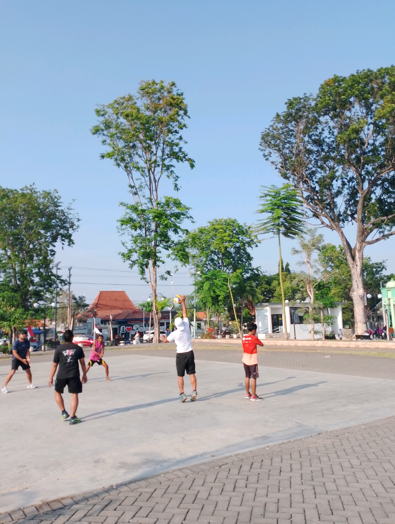 This image depicts an outdoor scene on a paved court, likely a sports facility, in Probolinggo, Indonesia. The sun is shining, casting distinct shadows on the ground, indicating it is daytime. Several individuals are engaged in a game of volleyball. One person, wearing a white shirt and black shorts, is in the process of serving the ball. Another individual, in a pink top and black shorts, is positioned to receive. Two other individuals are also present on the court, one in a dark blue shirt and black shorts, and another in a black shirt with a graphic design and black shorts. In the background, there are trees, buildings with red tiled roofs, and a clear blue sky. A sign that reads "I LOVE PROBOLINGGO" is visible on one of the buildings. The overall environment suggests a public recreational area within the city.