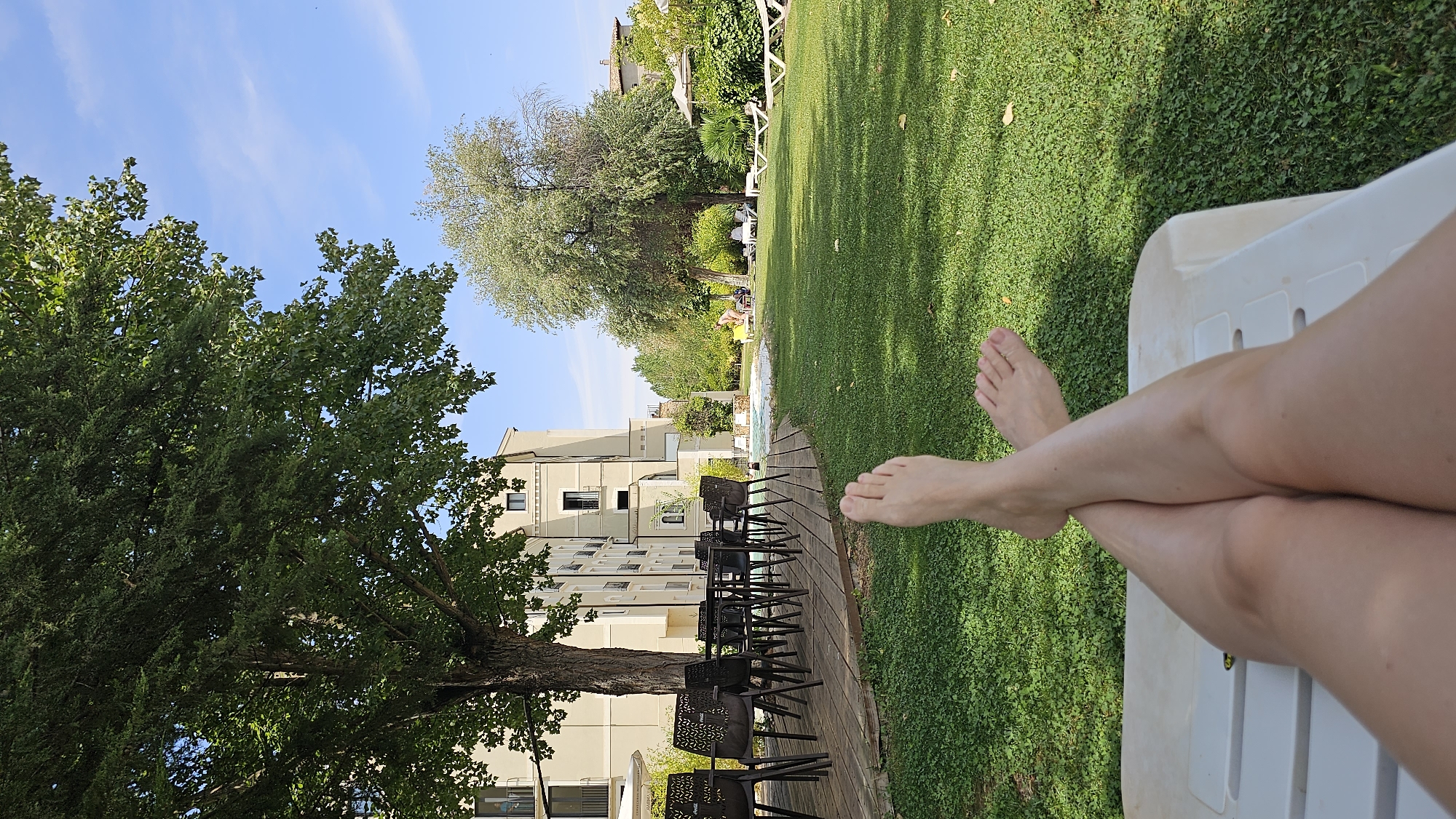 The image depicts a first-person perspective of someone relaxing on a white lounge chair in a sun-drenched outdoor area, likely a hotel or resort. The person's bare legs and feet are crossed at the ankles and are in the foreground, resting on the lounge chair. The background features a lush green lawn, a wooden deck with tables and chairs, a swimming pool, and a multi-story building. There are also several trees and some lounge chairs with umbrellas visible on the lawn. In the distance, there are people by the pool area. The sky is clear and blue with a few wispy clouds. The scene suggests a peaceful and leisurely atmosphere.