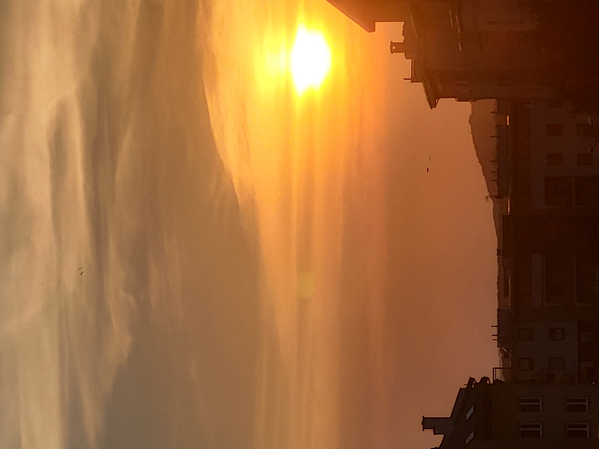 The image depicts a sunset over a city. The sun is a bright, glowing orb in the upper right quadrant, casting a warm orange and yellow light across the sky. Wispy clouds streak across the sky, tinged with the colors of the sunset. Silhouetted against the bright sky are several buildings, their rooftops and windows dark and indistinct. In the distance, a faint outline of hills can be seen. Two small, dark specks, possibly birds, are visible in the sky. The overall mood is serene and peaceful, capturing the beauty of a city at dusk.