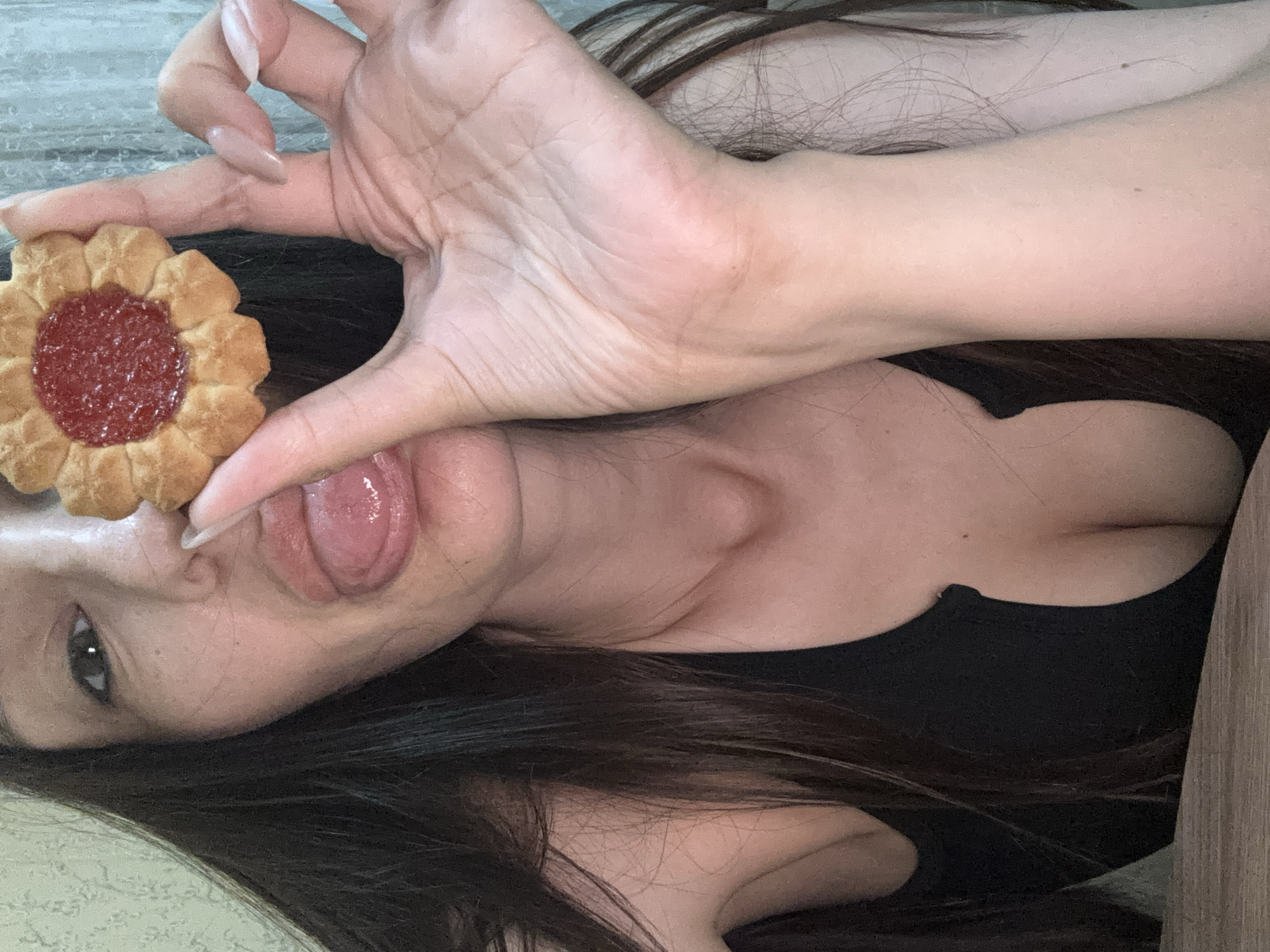 The image displays a close-up of a person, likely female, lying down, with the frame rotated such that her head is to the left. Her long, dark hair is spread across the background and around her shoulders. The central activity shows her playfully interacting with a single, flower-shaped cookie. Her right hand, featuring long, light-colored (possibly nude-pink) fingernails, holds the cookie directly in front of her face, while her tongue is extended out of her mouth towards the cookie, suggesting a fun or teasing gesture.
Visible details include her right eye looking towards the viewer, a portion of her nose, and her mouth. She is wearing a black top or bra, part of which is visible on her chest. The cookie is light golden-brown with a distinctive fluted or scalloped edge, and its center is filled with a bright red, jelly-like substance, characteristic of a jam cookie. The background is indistinct, appearing as a light-colored surface. The overall mood conveyed is playful and lighthearted. This scene is set in Sochi, Russia. There is no visible text in the image.