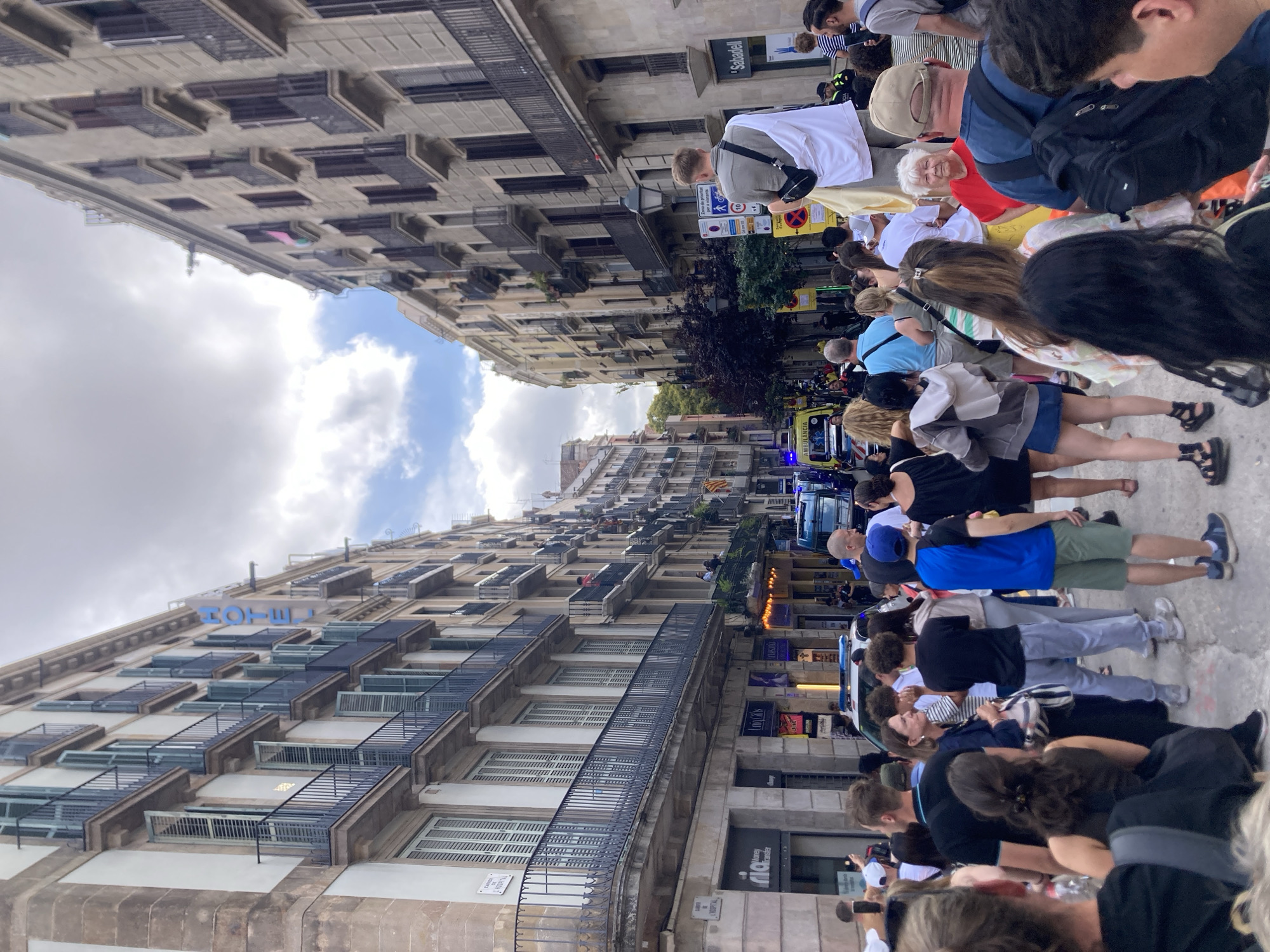 The image depicts a street scene in Barcelona, Spain, with a crowd of people gathered on a wide avenue lined with tall, historic buildings. The architecture features balconies with black railings and numerous windows, some with closed shutters. A prominent blue sign on one of the buildings reads "HOTEL".
In the foreground, a diverse group of people are visible from behind, facing away from the camera, suggesting they are observing something further down the street. Some individuals are holding up mobile phones, likely to take pictures or videos.
Further down the street, several vehicles are present, including what appears to be an ambulance with its emergency lights activated, and a police car. The presence of these emergency vehicles indicates a possible event or situation that has drawn the attention of the public and authorities.
The street is paved, and there are streetlights and signs visible, typical of an urban environment. The sky is partly cloudy, suggesting moderate weather conditions. The overall impression is of a busy public space in Barcelona, possibly during a daytime event or gathering.