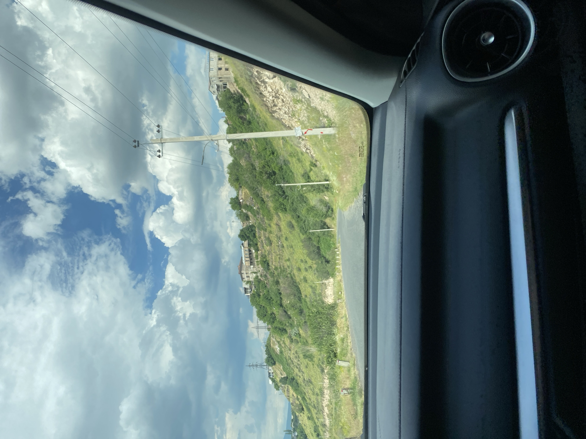 The image shows a view from inside a car looking out at a winding road in the city of Ptghni, Armenia. The road curves up a grassy hillside dotted with trees and houses. The sky is partly cloudy with bright blue patches visible. Power lines run along the side of the road, supported by utility poles. The car's dashboard is visible in the foreground, with a circular air vent on the right side. The overall scene depicts a sunny day in a semi-urban environment with a hilly landscape.