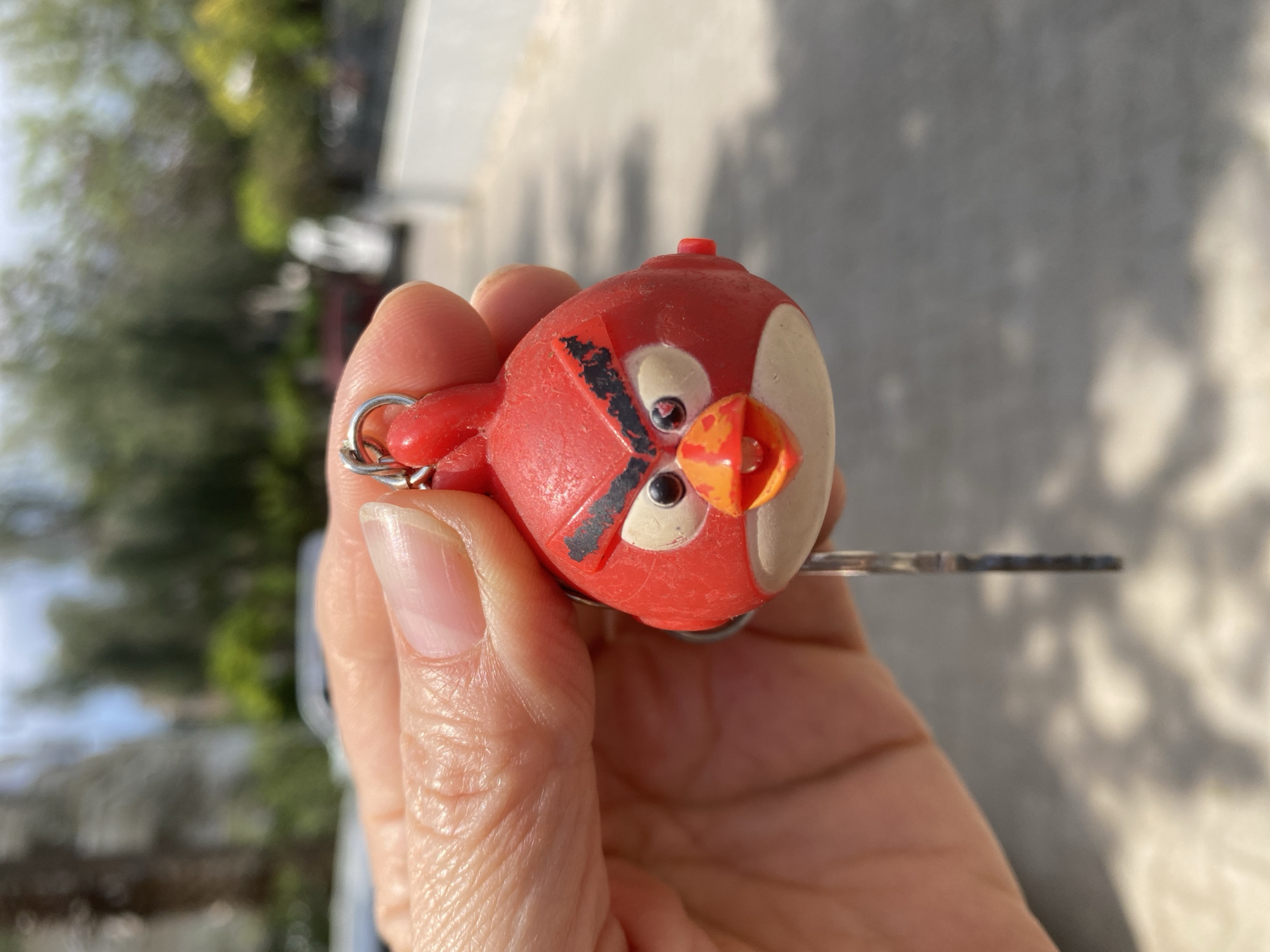 A close-up image shows a hand holding a keychain featuring an Angry Birds character. The hand belongs to an individual with a light skin tone. The keychain is attached to a key. The background is blurred, but suggests an outdoor setting with trees and buildings. Without additional information, pinpointing the location specifically to Alanya, Turkey, is not possible from visual analysis alone.