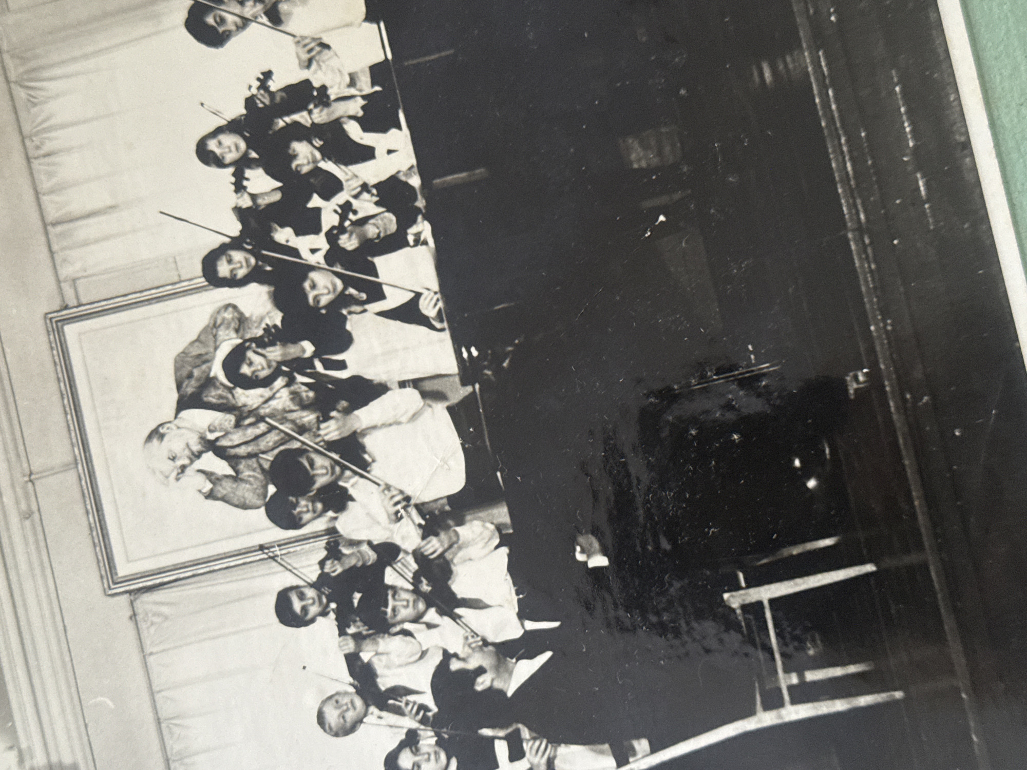 A black and white photograph shows an ensemble of people, seemingly musicians, playing stringed instruments, likely violins. They are dressed in formal attire, with some wearing white shirts and dark jackets or vests. The setting appears to be indoors, as suggested by the framed picture on the wall to the left and the dark area to the right containing what looks like furniture or stage props. The photograph is of a performance or rehearsal. The city of Ramat Gan, Israel cannot be determined from the image.