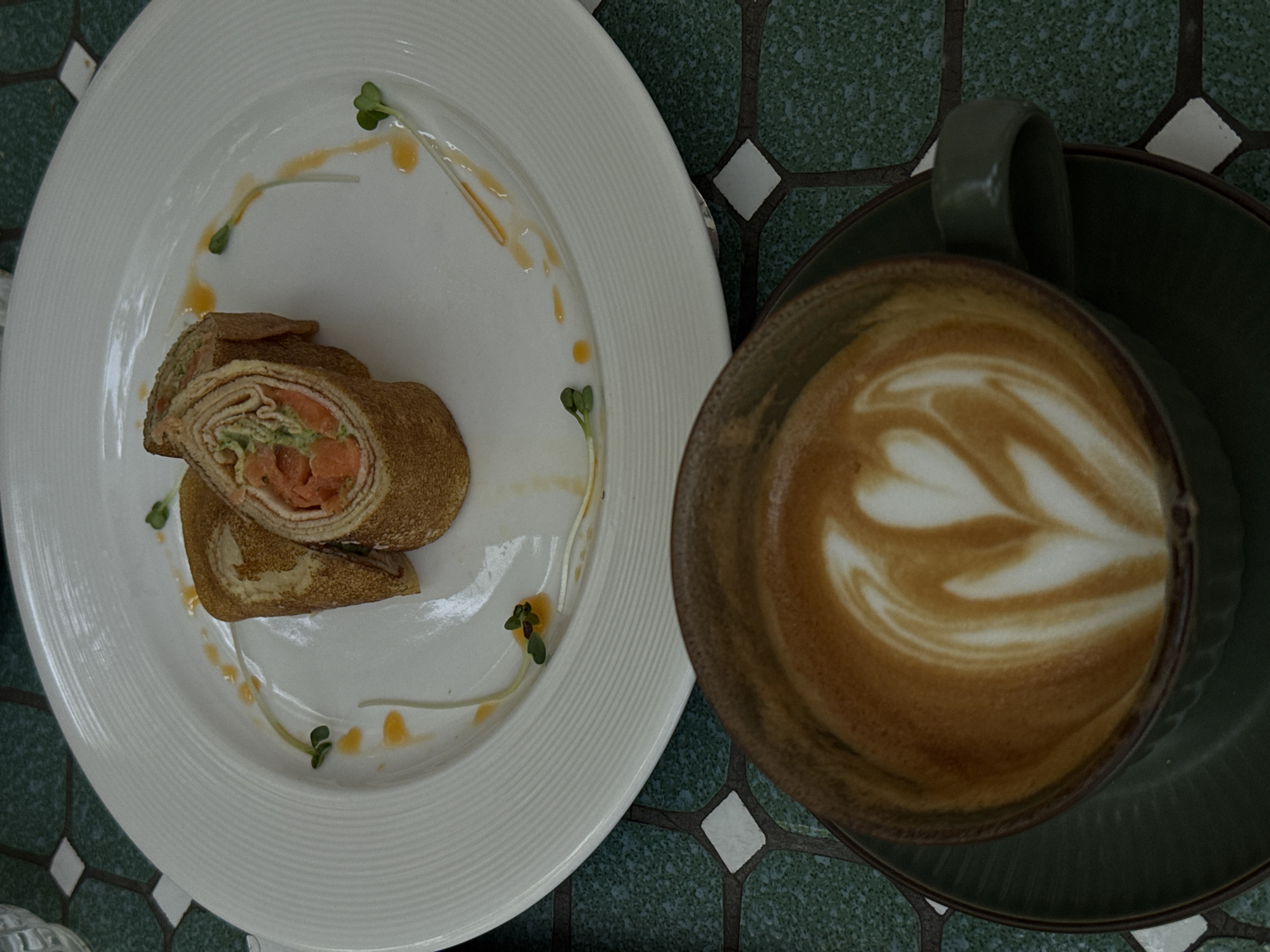 The image shows a top-down view of a plate and a coffee cup, likely taken in a café setting in an unknown city.
The plate, white with a ribbed edge, holds what appears to be a rolled crepe or wrap cut into two pieces. The filling visible in the cut pieces includes salmon and some green vegetable, perhaps avocado or sprouts. The plate is decorated with a drizzle of sauce and small sprigs of greenery.
Beside the plate is a dark green coffee cup and saucer. The coffee has latte art with a heart or leaf pattern on its surface.
The table beneath the plate and cup has a tiled surface with a green and white geometric pattern. The overall impression is that of a light meal or snack with coffee.