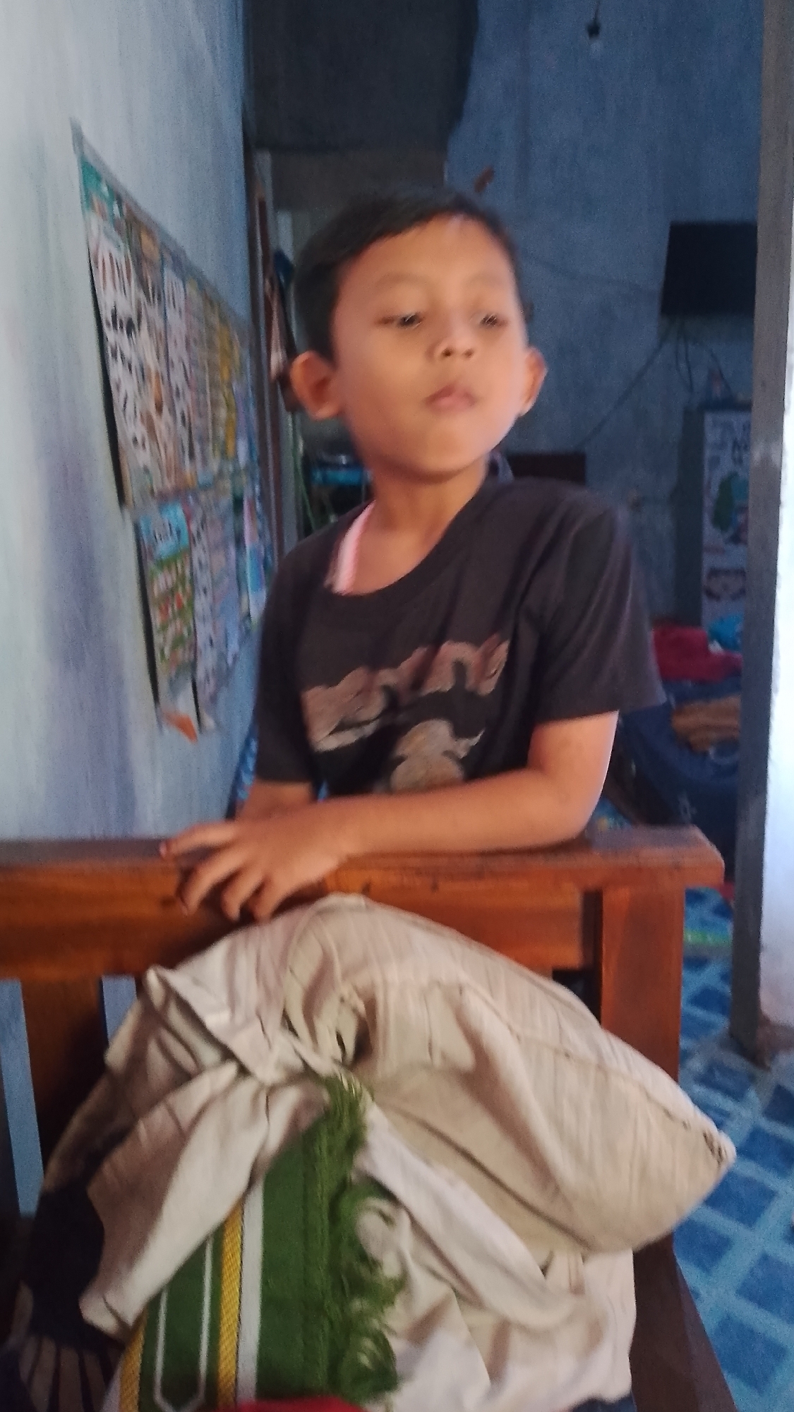 The image depicts a young boy, appearing to be of Southeast Asian descent, seated in a wooden chair in what is likely a residential setting. The boy is wearing a dark t-shirt with a graphic design and a light-colored undergarment. His hands are resting on the edge of the chair. In front of him, a light-colored pillow with a patterned fabric is placed on the chair. The background is dimly lit, showing a wall with some posters or artwork and a glimpse of what appears to be a doorway or opening. The flooring is tiled in a blue pattern. The overall environment suggests a humble, indoor domestic space. The image is of Indonesian origin, with the location specified as Karanglangu, Indonesia.