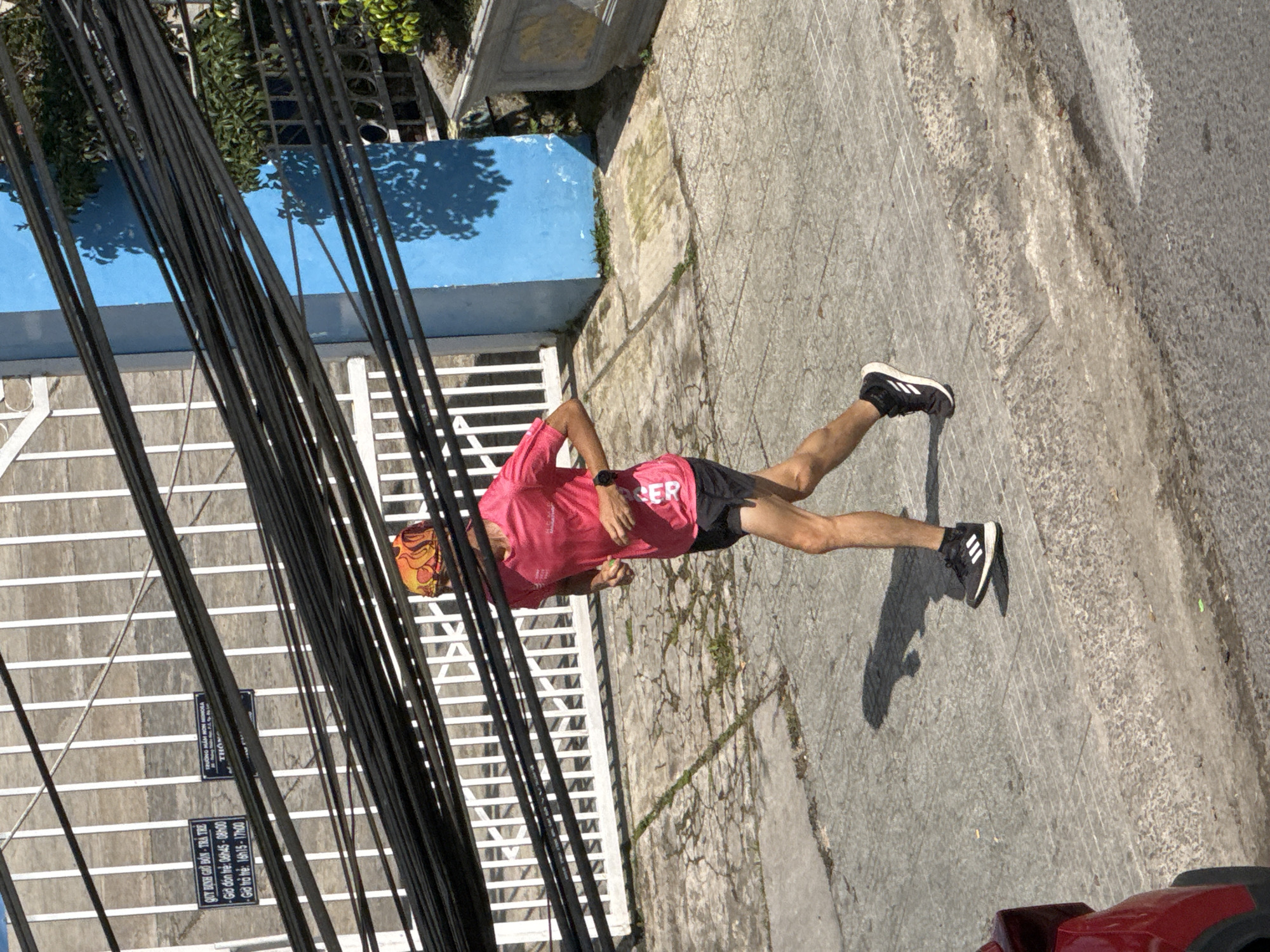 A person wearing a bright pink t-shirt, black shorts, a colorful bandana, and black athletic shoes is jogging on a paved sidewalk in Da Lat, Vietnam. The runner has a watch on their left wrist. The sidewalk is cracked and uneven, with some weeds growing through the cracks. Behind the runner is a white fence and a building with a bright blue wall. A dense cluster of black electrical wires hangs low over the scene. In the foreground, the rear of a red vehicle is visible on the left side of the image. The sun casts a strong shadow of the runner on the sidewalk. Two signs are attached to the wall behind the fence; one sign appears to be a schedule for a daycare or kindergarten with times listed in Vietnamese.
