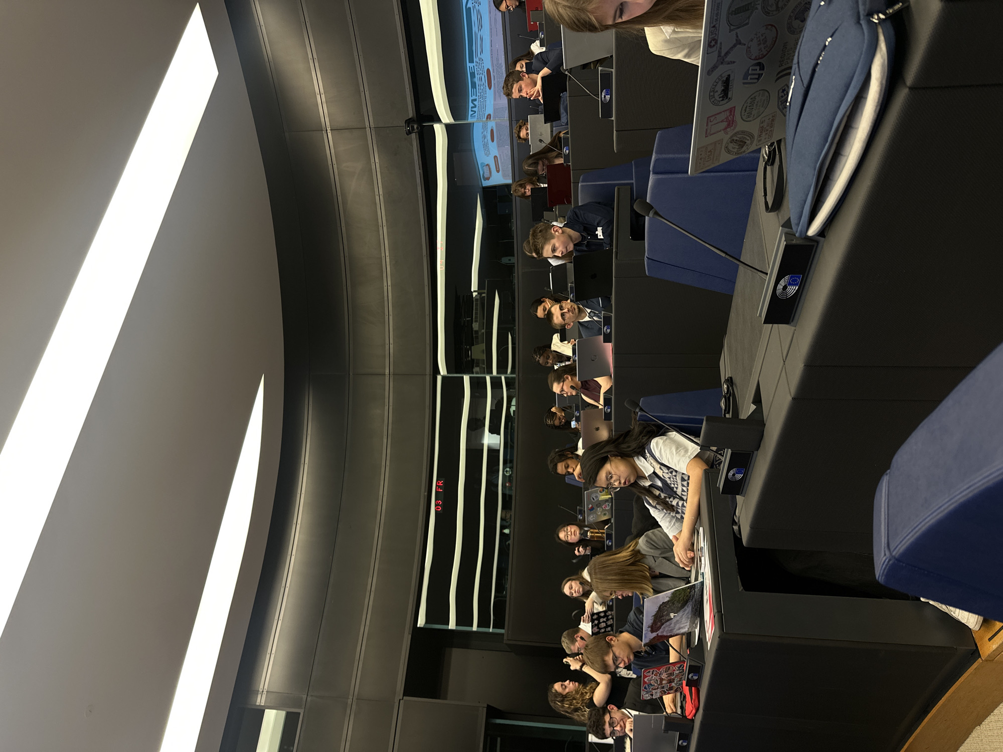 The image shows an interior view of the European Parliament in Strasbourg, France. Multiple rows of seated individuals are present, each person appearing to be working with a laptop or engaging with materials on the desk in front of them. Microphones are visible at each workstation. A screen displaying information is visible. The architecture consists of curved lines, dark panels, and light-emitting fixtures on the ceiling. The scene depicts a structured environment, likely during a parliamentary session.
