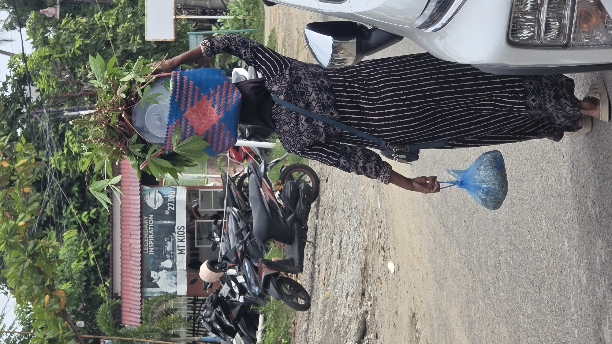 A woman in a patterned long-sleeved top and striped dress stands on a paved road in Lhokseumawe, Indonesia. She holds a blue plastic bag full of small fish in her right hand. On her head, she carries a woven basket filled with leafy greens. A white car is parked on the right side of the image, and several motorcycles are parked in front of a building on the left with a sign that reads "MT KIOS LEGENDARY INSPIRATION." The background includes trees and foliage.