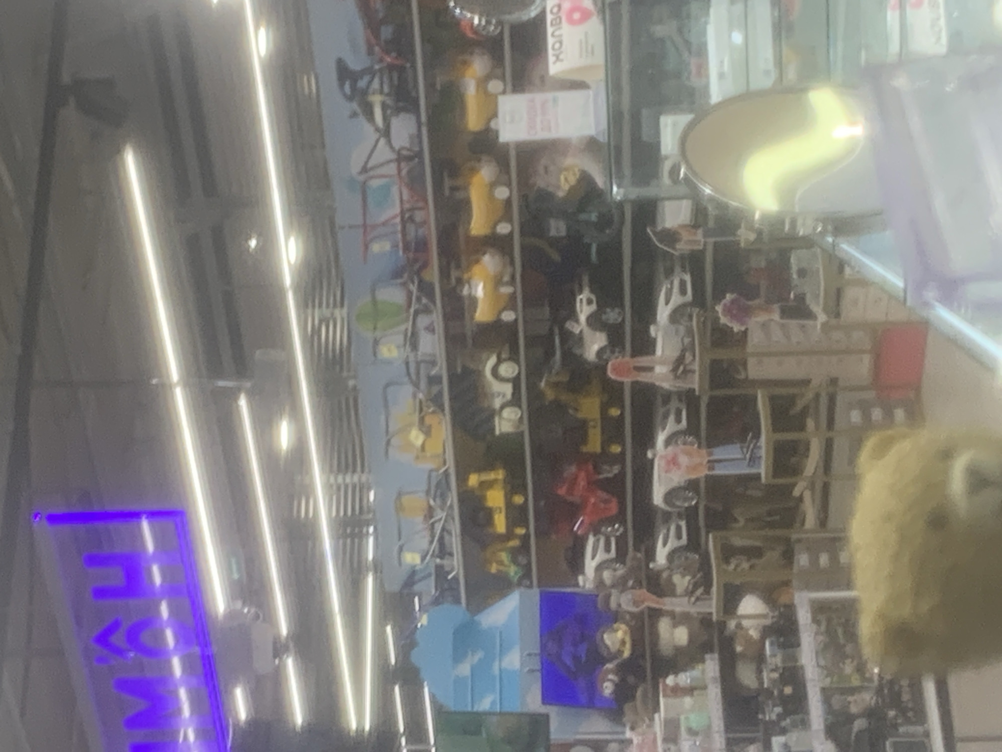 The image displays an indoor retail environment, likely an aisle within a large store such as a hypermarket or toy store. The image is rotated 90 degrees clockwise from its natural upright orientation.
**Subjects:** The primary subjects are various products displayed on shelves. These include numerous toy vehicles, notably yellow construction vehicles (such as diggers or loaders), red cars, and white cars or trucks, suggesting a children's or toy section. In the immediate foreground on the right side (when rotated), a blurry, yellowish-brown plush toy is visible. The shelves behind are stocked with various other, less discernible items. No people are clearly visible.
**Activity or event:** The image captures a static scene of a store aisle; no specific activity or event is taking place.
**Notable details:** The store is brightly illuminated by long, linear fluorescent or LED strip lights on the ceiling, which create distinct reflections on the floor and other surfaces, indicating an indoor commercial setting. A circular or oval reflective surface (possibly a mirror or display element) is visible in the upper central area (when rotated), showing a wavy reflection of light. The overall color palette is somewhat muted, with prominent yellows and reds from the toys and a bright purple from a neon sign.
**Visible text:**
* **Left side (bottom if upright) of the rotated image:** A vertical purple neon sign displays characters that appear to read "<MOH" when viewed from bottom to top.
* **Upper right side (top if upright) of the rotated image:** A pink rectangular sign with white Cyrillic text is partially visible, potentially reading "ХОЛВО" or "ХОЛВА" (a type of sweet common in Russia). Below it, another white sign with dark, indistinct text can be seen.
