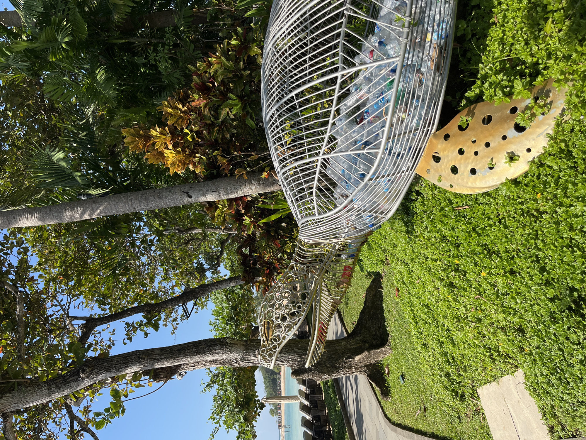 This is a sunny, outdoor scene in Kuta Selatan, Indonesia, depicting a metal turtle sculpture filled with plastic bottles. The sculpture is placed on a lush green lawn, with a stone pathway curving to its left. Behind the sculpture, a tropical garden with vibrant green and yellow foliage is visible, bordered by palm trees and other leafy vegetation. Beyond the garden, lounge chairs and umbrellas are set up on a beach overlooking a calm, turquoise ocean under a clear blue sky. The turtle sculpture is intricately made of white metal bars, forming its body and head, with its mouth open, revealing the collection of plastic water bottles inside. A golden, leaf-shaped piece with circular perforations lies on the grass next to the turtle's shell. The environment suggests a resort or public area near a beach.