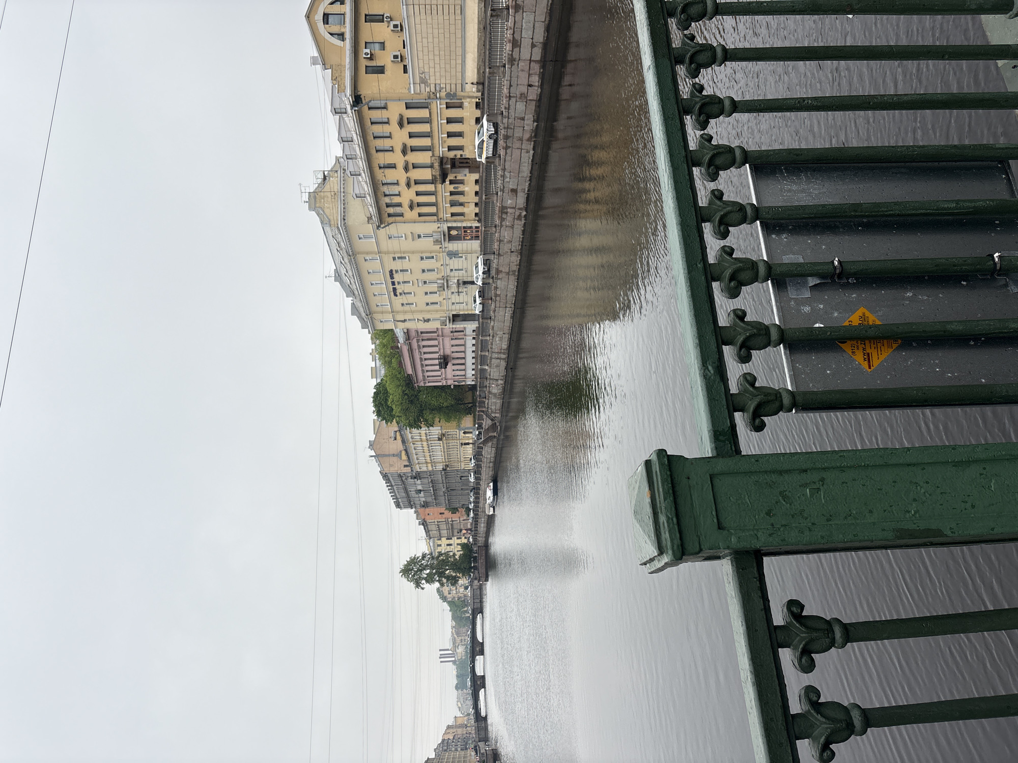 This image showcases a canal in Saint Petersburg, Russia. The viewpoint is from a bridge, with a green, decorative metal railing visible in the foreground. The canal runs alongside buildings, some of which are painted in muted colors like yellow and pink. Cars are parked along the canal's edge. The sky is overcast, suggesting a cloudy or rainy day. The water in the canal is still, reflecting the buildings and sky. In the background, there is another bridge spanning the canal. Powerlines are visible in the sky.