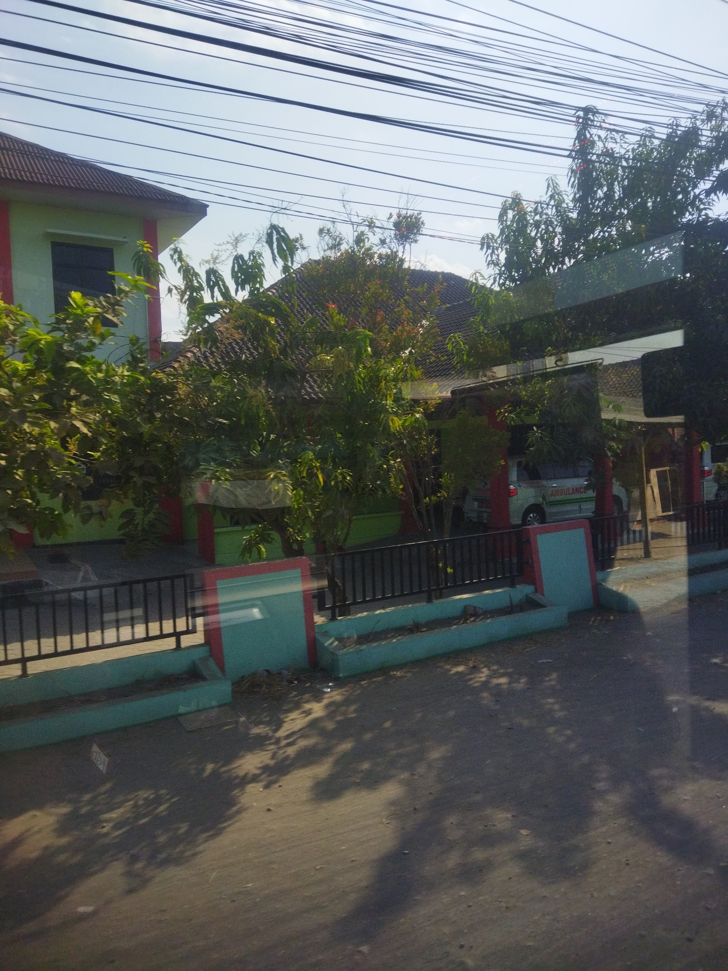 The image captures a street-level view of a building in Nguter, Indonesia, characterized by its light green facade with red accents. The building's roof is tiled with dark brown shingles, and a dark-framed window is visible on the left side. Vegetation, including trees with green leaves, surrounds the building and partially obscures its view. A dark metal fence runs along the front of the property, adjacent to a low, teal-colored wall with red trim. The wall appears to be a planter box. A white minivan with "AMBULANCE" written in green lettering is parked under a covered area to the right of the building. The ground in front of the building is paved, with shadows of trees cast across it, suggesting it is daytime. Overhead, multiple power lines crisscross the sky, which is a clear blue.