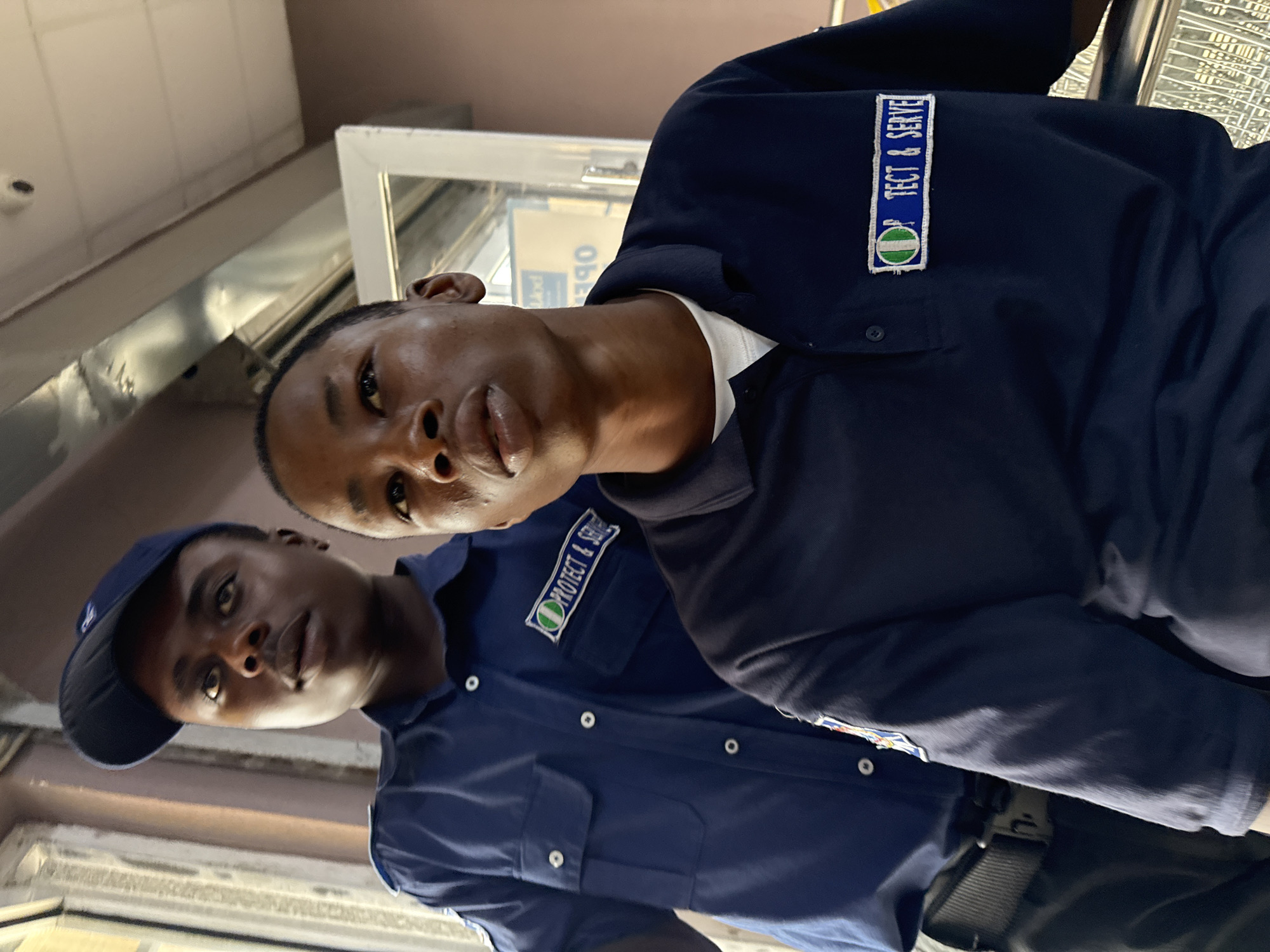 Two men are indoors. Both are wearing blue uniforms bearing the insignia "PROTECT & SERVE". One man wears a blue baseball cap. Location is presumed to be Ado, Nigeria.