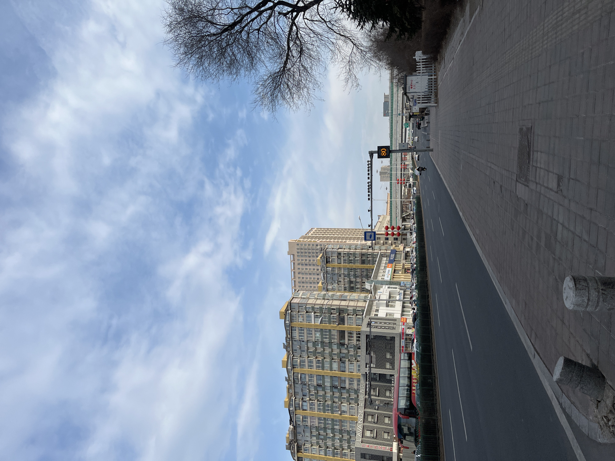 The image depicts a street scene in Changchun City, China. A multilane road stretches into the distance, flanked by tall buildings on one side and a wide sidewalk on the other. The buildings appear to be a mix of residential and commercial properties, with several stories and varying architectural styles. On the street, cars and a bus are visible, moving in the same direction. A traffic sign displays the number "80," likely indicating a speed limit. The sky is partly cloudy. A bare tree branch is present in the top left corner of the frame.