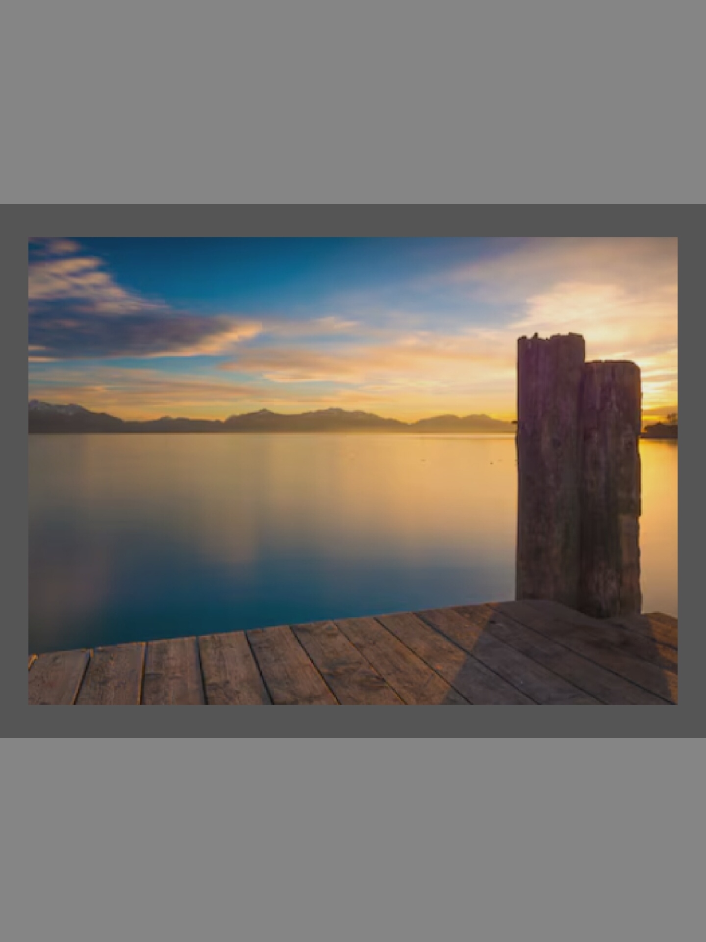 The image captures a serene sunset or sunrise scene over a large body of water, likely a lake, with mountains silhouetted in the distance. The sky is a dramatic display of warm colors, transitioning from golden yellows and oranges near the horizon to soft blues higher up, with scattered clouds reflecting the vibrant light.
In the foreground, a wooden pier extends into the lower right of the frame, its planks showing textured detail. At the end of the visible pier, two rough, weathered wooden posts stand tall, breaking the smooth horizon line. The water is calm and still, reflecting the warm hues of the sky and appearing glassy, with a gentle transition from golden tones near the horizon to deeper blues closer to the foreground.
The distant mountains are dark and indistinct against the bright sky, adding depth to the expansive landscape. No people or specific activities are visible, emphasizing the tranquility and natural beauty of the scene. The time of day is clearly dawn or dusk, characterized by the low, soft light and rich color palette. The weather appears clear and calm. There is no visible text.