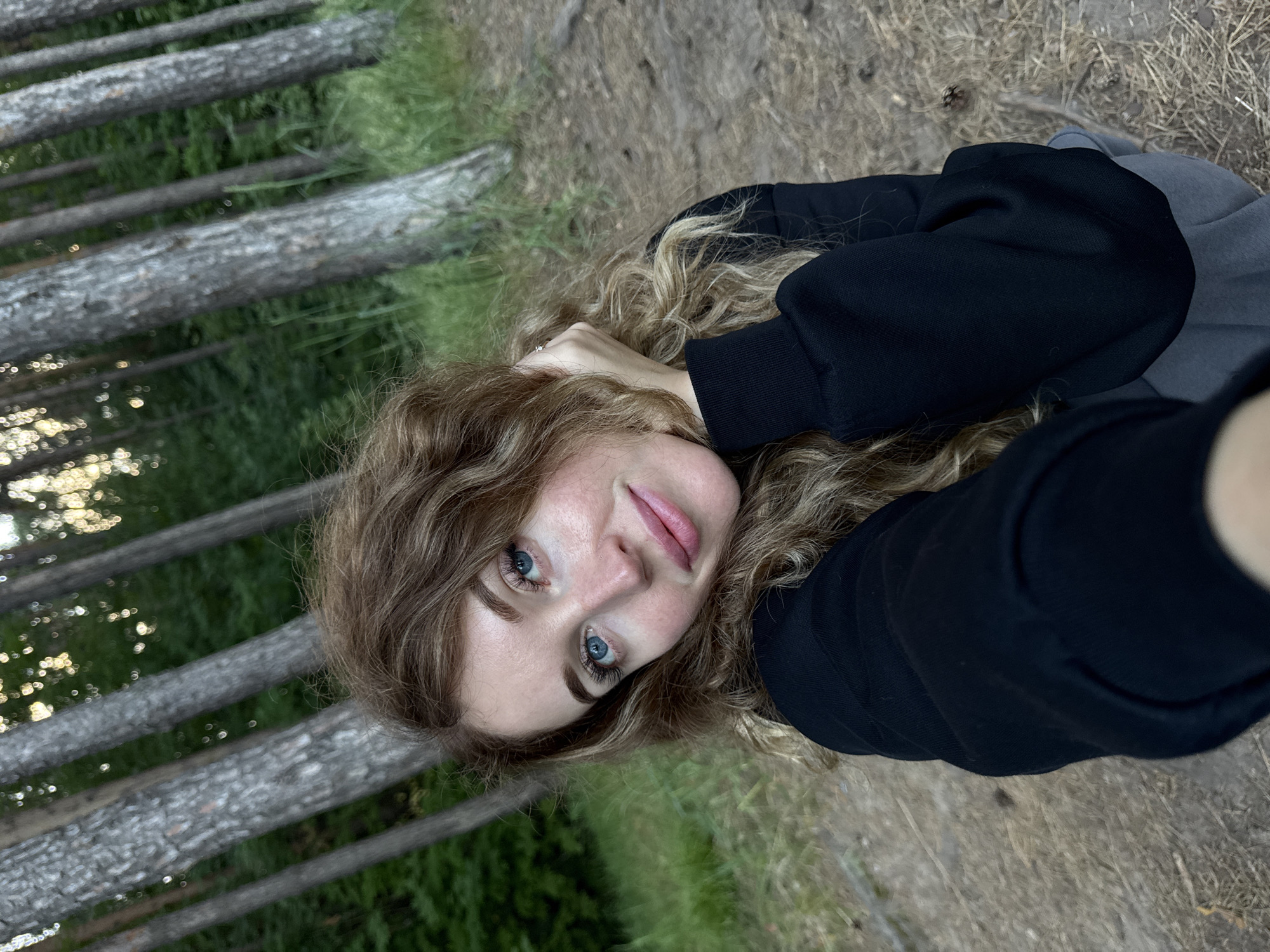 In a forest setting, a young Caucasian woman with long, curly blonde hair and blue eyes is pictured. She is wearing a black hooded sweatshirt and grey pants. Her left hand is resting on her head, and she is looking towards the camera with a slight smile. The background consists of tall, thin trees with green foliage. The ground is covered with pine needles and dry grass. The lighting suggests it is either early morning or late afternoon. The location is identified as Chaikovsky, Russia.
