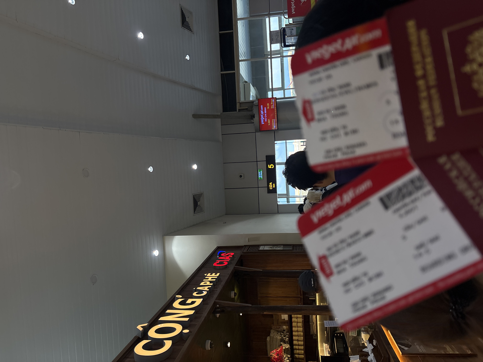 The photo shows a person holding a red passport and a Vietjet Air ticket at the Cam Ranh International Airport, located in Bac Cam Ranh Ward, Vietnam. The person is likely waiting in an airport terminal. A Cong Caphe coffee shop is visible in the background, as well as a VietjetAir sign indicating boarding gate 5.
