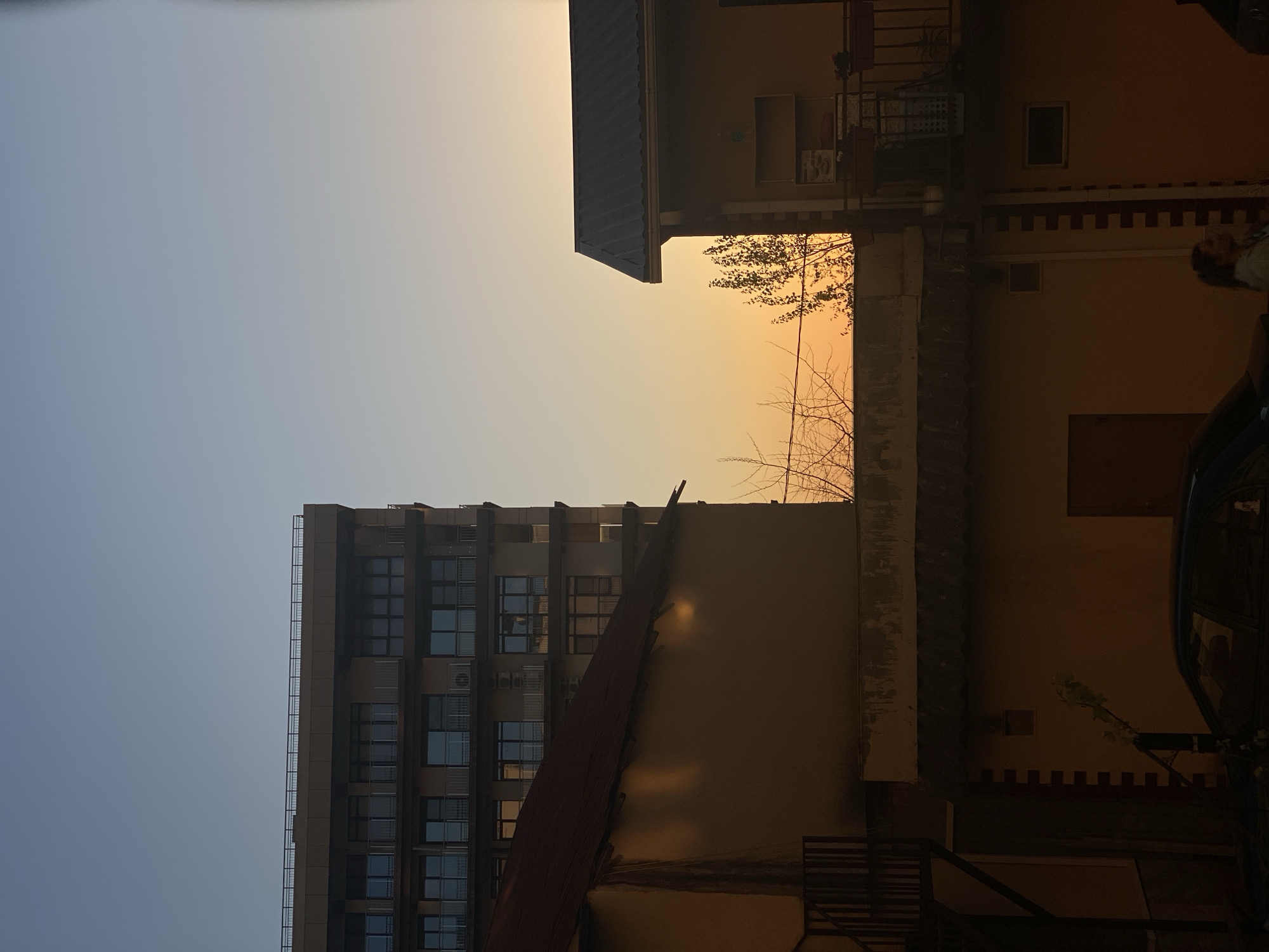 An urban scene at what appears to be sunset or sunrise, given the gradient sky from light blue to a warm orange and yellow hue. The image is taken from a low angle, looking up at several buildings.
On the left side of the frame, a tall, modern multi-story building with numerous windows, possibly an apartment or office complex, stands against the lighter blue portion of the sky. Its facade is dark, contrasting with the bright sky.
In the mid-ground, a lower, rectangular building with a dark, sloping roof is prominent. Its side wall, facing the camera, is a muted beige or light brown, catching some of the warm light from the sky. Bare tree branches are silhouetted against the orange sky behind this building.
To the right, a different building with a dark, gabled roof and light yellow-brown walls is visible. It features balconies or railings on its upper levels. A person's head and shoulders, wearing a light top, are partially visible on the far right edge, seemingly observing the scene. Below them, a section of what appears to be a dark vehicle is faintly seen. The lower wall of this building has a decorative pattern near its base.
The overall atmosphere is tranquil, with the warm, soft light of the sun creating subtle reflections on building surfaces and highlighting the silhouettes of structures and sparse tree branches. No discernible text is visible. The location context indicates Sochi, Russia.