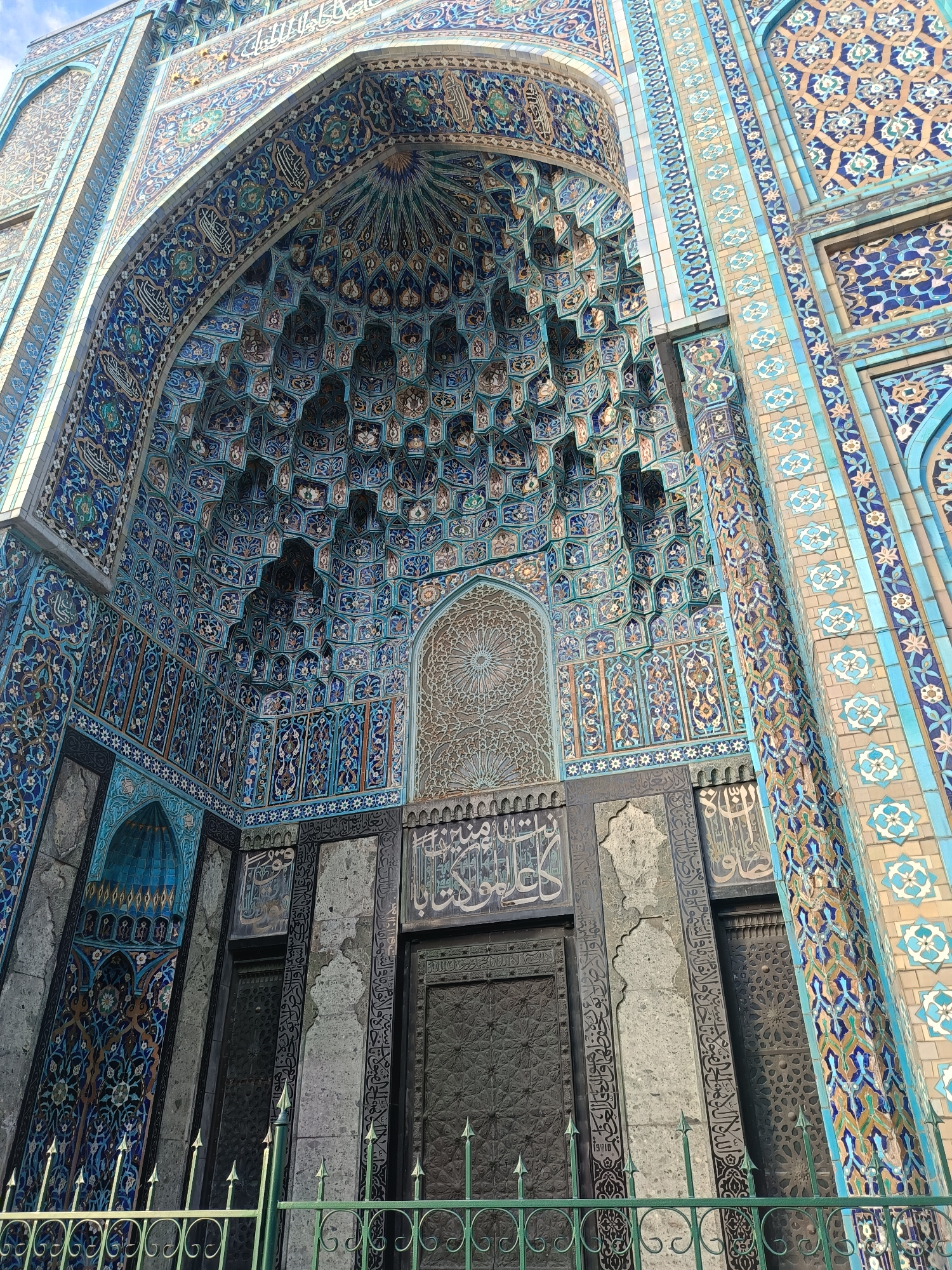 This is a low-angle, full-shot photograph of the exterior of the Saint Petersburg Mosque, located on the Petrograd Side of Saint Petersburg, Russia. The architectural facade is richly decorated with intricate blue and turquoise glazed tiles, forming complex geometric patterns and floral motifs. The main subject is a large, deeply recessed arched entryway, with a muqarnas (honeycomb vaulting) ceiling also covered in detailed tilework. Arabic calligraphy is visible on stone panels above two dark, ornate doors. A green metal fence with pointed finials runs along the bottom of the frame, in front of the mosque entrance. The sky is visible in the upper left corner. There are no people or animals in the image. The lighting suggests it is daytime.