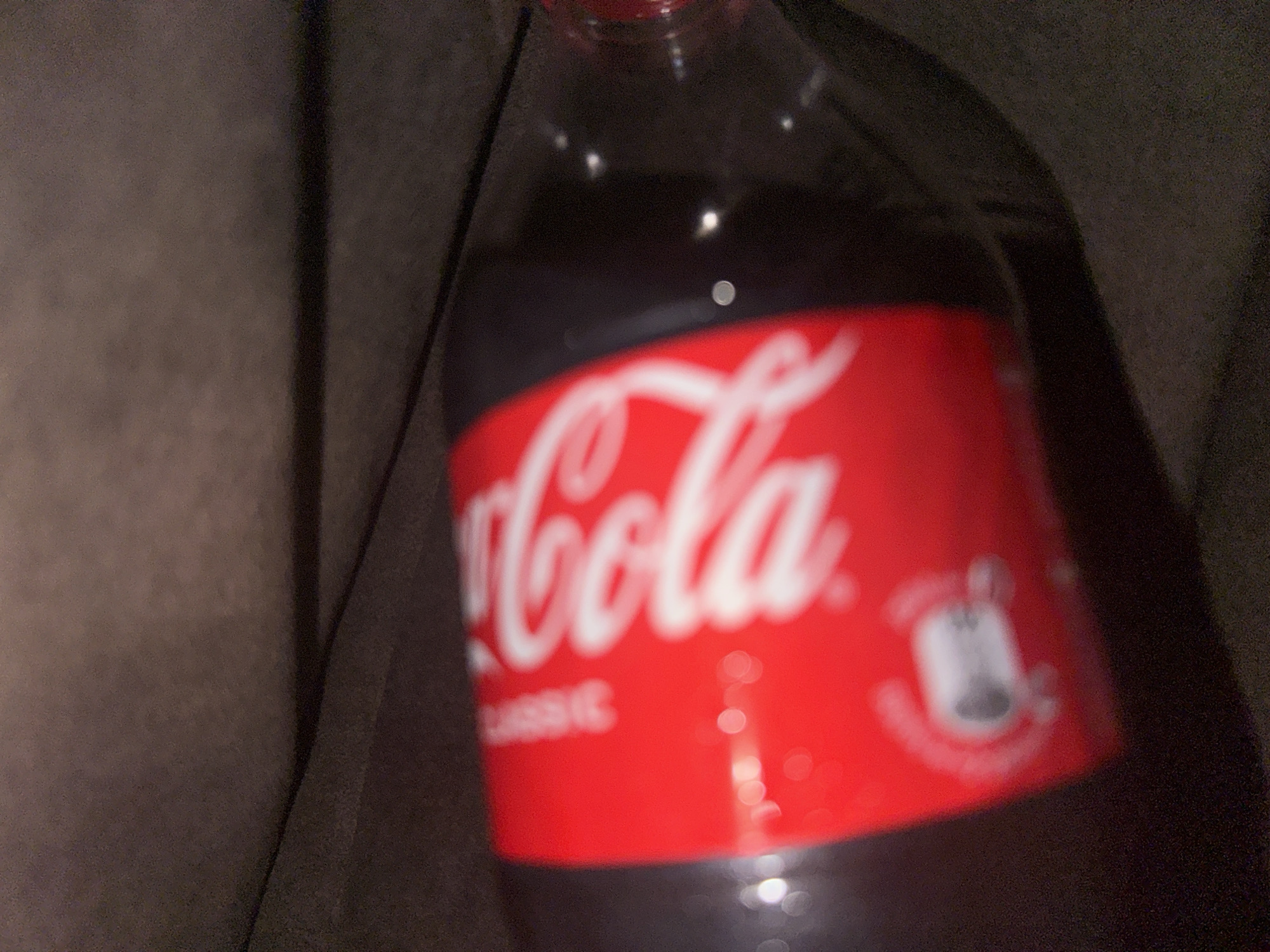 A close-up, slightly blurry image shows a partially visible bottle of Coca-Cola Classic. The red label with the white "Coca-Cola" script is prominently featured. The bottle is situated against a dark, neutral-toned background. The environment appears to be dimly lit and undefined. The location is Unknown, Unknown.