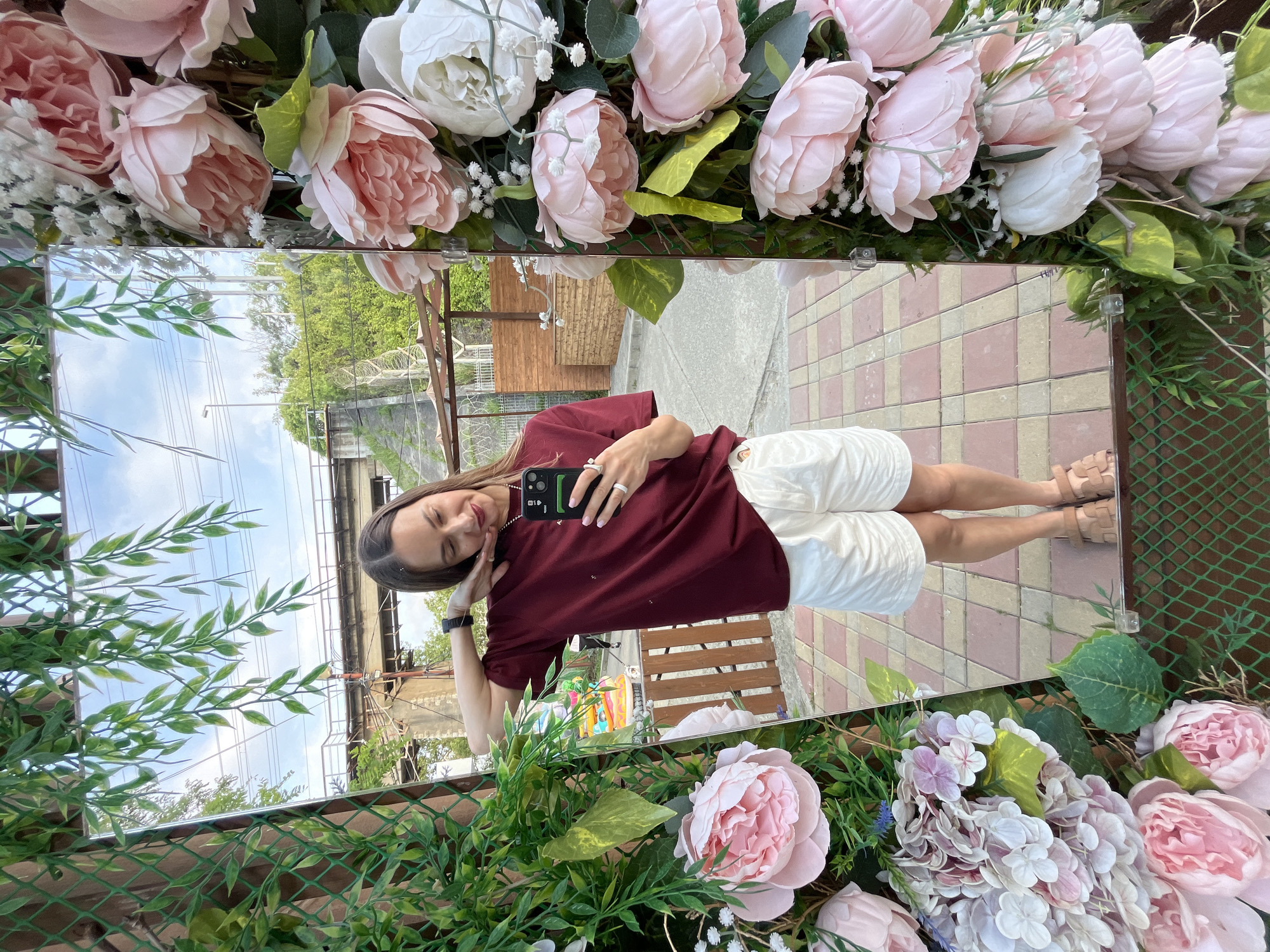 A young woman, with long brown hair and dark red lipstick, stands in front of a mirror, taking a selfie. She is wearing a maroon t-shirt and white shorts. Her right arm is bent, and her hand is resting on her cheek. The mirror reflects the woman and the surrounding environment, which appears to be an outdoor area decorated with artificial flowers and greenery. In the background, a pedestrian bridge can be seen, along with trees and buildings. The ground is paved with pink and beige tiles. The scene is likely located in Sochi, Russia, given the presence of a pedestrian bridge and the architectural style of the buildings.