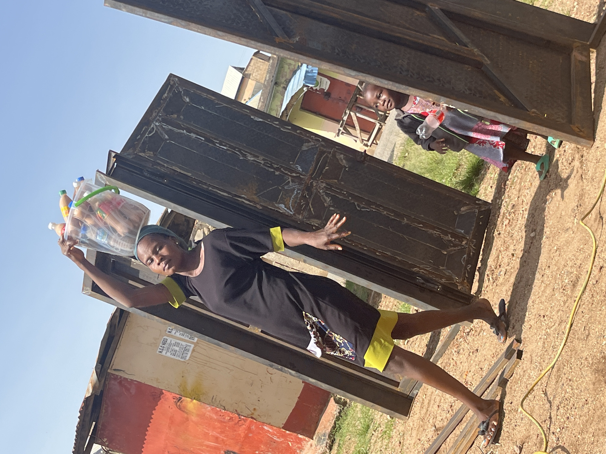 A woman, wearing a black dress with yellow trim and a headscarf, carries a clear plastic bag filled with bottles and bread on her head. She is standing and looking towards the camera. To her right, a young child in a patterned dress and dark jacket peeks out from behind a large metal gate. The child is holding a plastic bottle. The scene appears to be outdoors, with buildings and a blue sky visible in the background. The ground is sandy, and there is a yellow electrical cord running across it. The overall impression is of a bright, sunny day. The image is oriented vertically, with the subjects appearing sideways.
Visible text includes "N.G.O NEED" and what appears to be a phone number starting with "080". The location context is Jos, Nigeria.