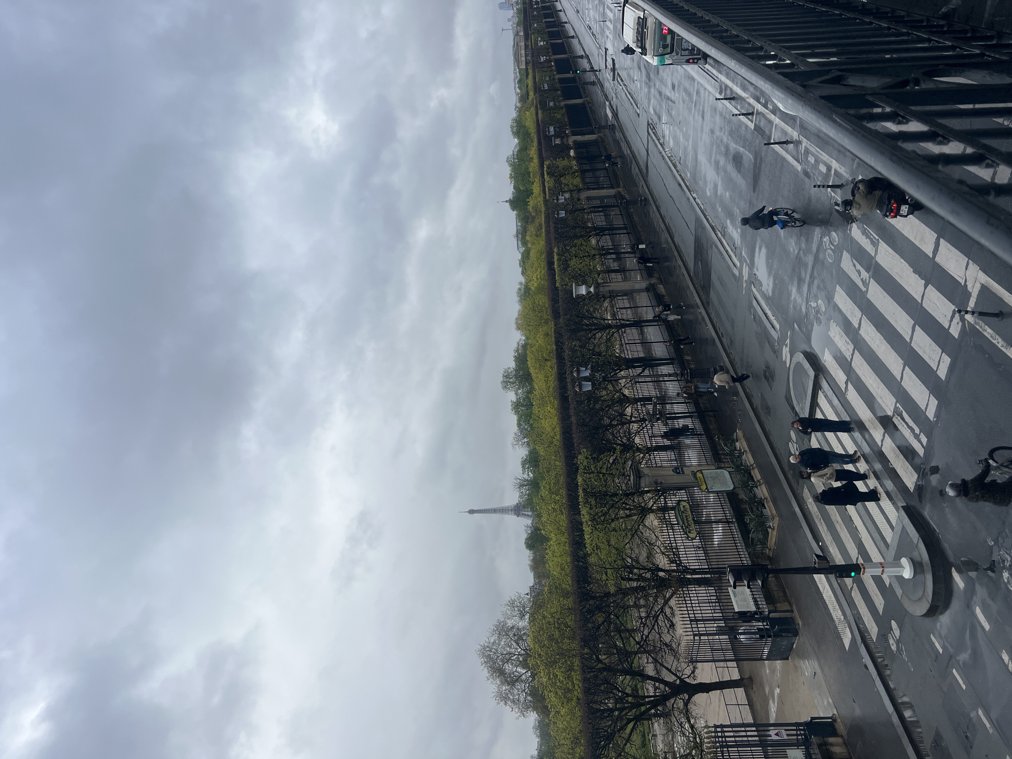 The image depicts an elevated view of a city street and an adjacent park in Paris, France, on an overcast day with wet asphalt indicating recent rain. The Eiffel Tower is visible in the distant background, partially obscured by a line of trees with varying degrees of foliage.
In the midground, a long, dark metal fence with decorative urns and numerous black lamp posts separates a park, likely the Tuileries Garden, from a wide, multi-lane street. The park features a dense arrangement of trees, some with bare branches and others showing fresh green leaves. A "METROPOLITAIN" sign, indicating a Paris Metro entrance, is affixed to a stone pillar by the fence on the left side of the street.
The street itself is dark and reflective from the moisture. A prominent white zebra crosswalk is visible in the lower-middle portion of the frame, with a central pedestrian island. A traffic light on this island displays a green signal. Four pedestrians are clustered on the central island of the crosswalk, three facing generally left and one facing right.
Vehicular and pedestrian activity is present:
- A person on a bicycle, wearing a dark jacket and helmet, rides away from the viewer in a designated lane beyond the crosswalk.
- A motor scooter with a rider in a helmet and a green jacket travels away from the viewer in the main vehicle lane, ahead of the crosswalk.
- A green city bus, marked with the number 72, is partially visible on the far right, moving away from the viewer.
- Another cyclist in a dark jacket and light helmet is at the far left edge of the frame, heading towards the viewer.
- Several individuals are visible walking on the sidewalk alongside the park fence.
- Black traffic bollards are spaced along the street, delineating lanes and guiding traffic.
- Bicycle lane markings are faintly discernible on the wet street surface.
The upper third of the image is dominated by a uniformly grey, cloudy sky. The perspective is from an elevated position, with a dark metal railing partially visible in the lower right foreground.