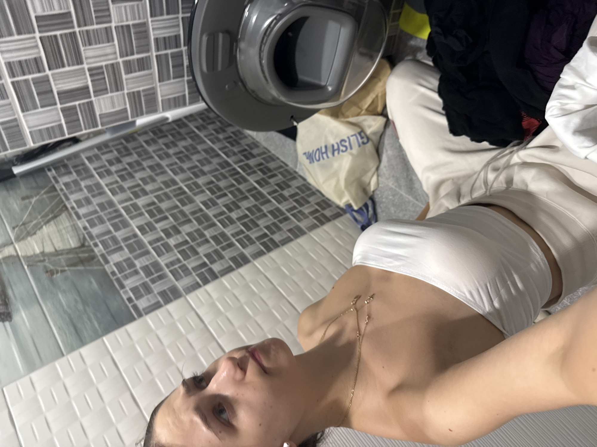 A young woman is sitting on the floor next to a washing machine in what appears to be a laundry room or bathroom. She is wearing a white strapless top and light-colored pants. Her hair is dark and pulled back. She is looking off to the side with a thoughtful expression. There are clothes on the floor, some in a laundry bag and others piled around the washing machine. The walls are tiled with a mosaic pattern. The lighting is artificial and seems to be coming from overhead. The scene is domestic and suggests a person is in the process of doing laundry. The specific location in the city of Sofia, Bulgaria cannot be determined from the provided image.
