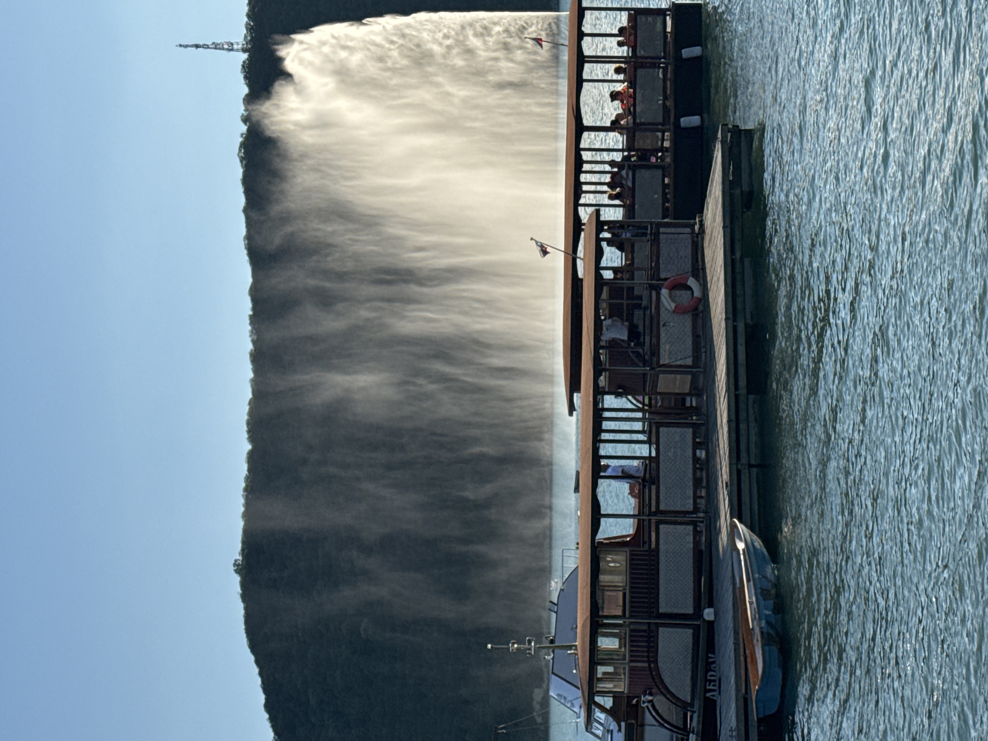 In the city of Abrau-Dyurso, Russia, two tour boats are docked at a wooden pier on a body of water. The boats are filled with passengers, many of whom are wearing life vests. In the background, a massive man-made fountain is spraying water high into the air against a dark, tree-covered hillside. The sky is clear and blue. A radio tower is visible on the crest of the hill. The scene appears to be a popular tourist attraction.