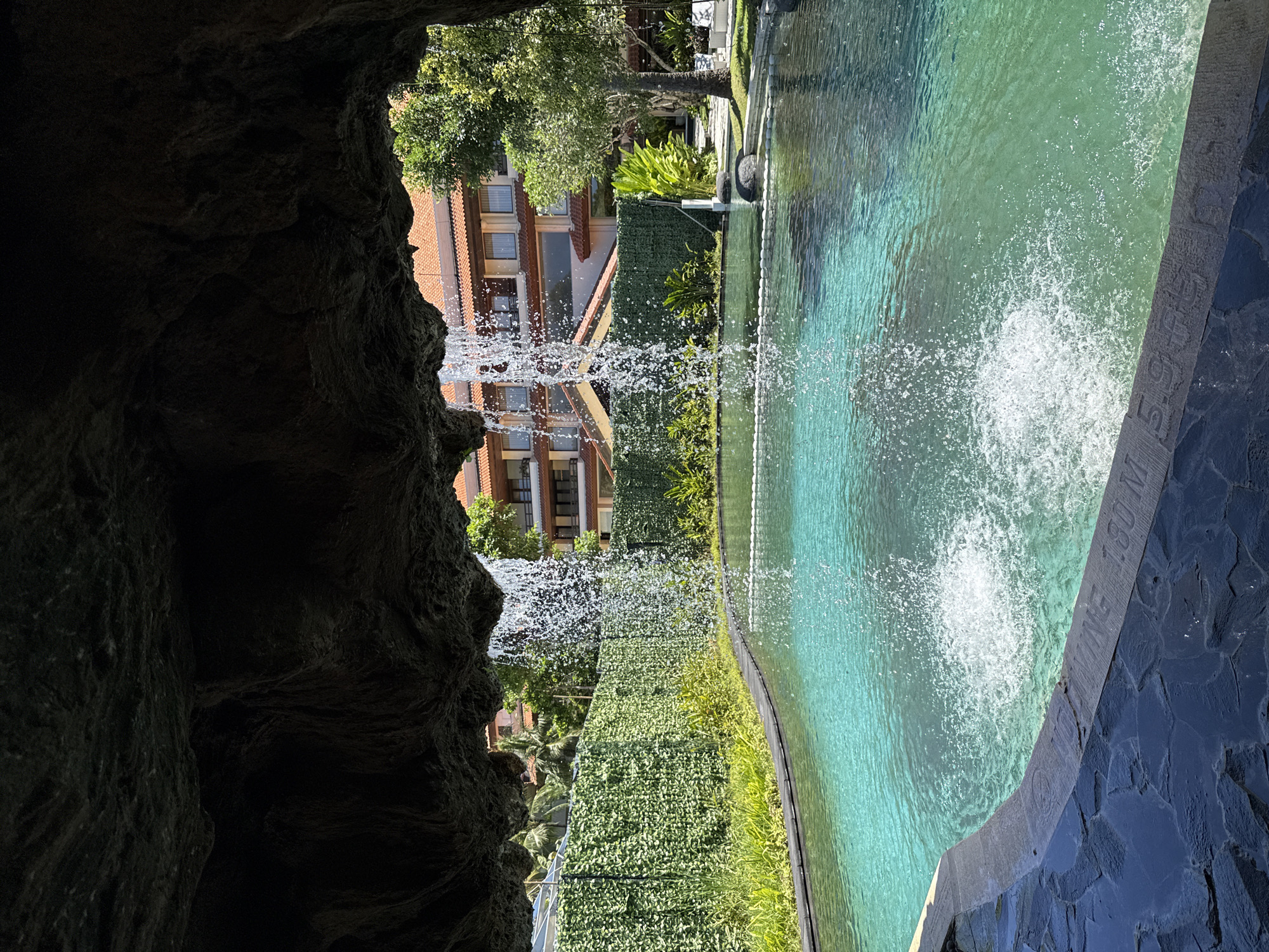 Captured from within a dark, rocky cave-like structure, the image offers a view into a scene in Nusa Dua, Indonesia. The main subject is a vibrant, turquoise-colored swimming pool featuring multiple fountains that spray water upwards, creating a sparkling effect. Adjacent to the pool, there is a vertical green wall. Beyond this are a building with reddish-brown walls and windows. The pool's edge is marked by a stone border with what appears to be lettering engraved. The environment is sunny and appears to be tropical.
