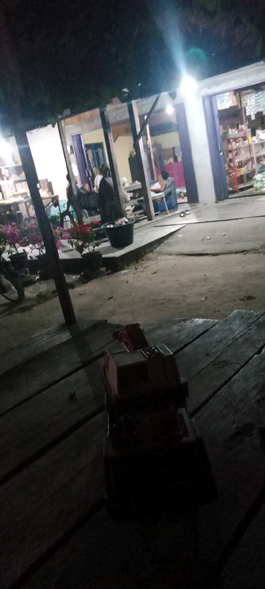 The image depicts an outdoor scene at night in what appears to be a market or shop area in Kerumutan, Indonesia. In the foreground, a red toy truck is placed on a wooden surface. Behind the toy, a paved area is visible, leading to a building with several individuals present. One person is seated at a table, and another is standing nearby. Shelves stocked with various items are visible inside the building, suggesting it is a store. Potted plants, including pink and red flowers, are placed along a raised concrete area in front of the building. The overall lighting suggests the presence of artificial illumination from within the building and possibly streetlights. The ground surface appears to be a mixture of dirt and pavement, with some puddles indicating recent rain or moisture.