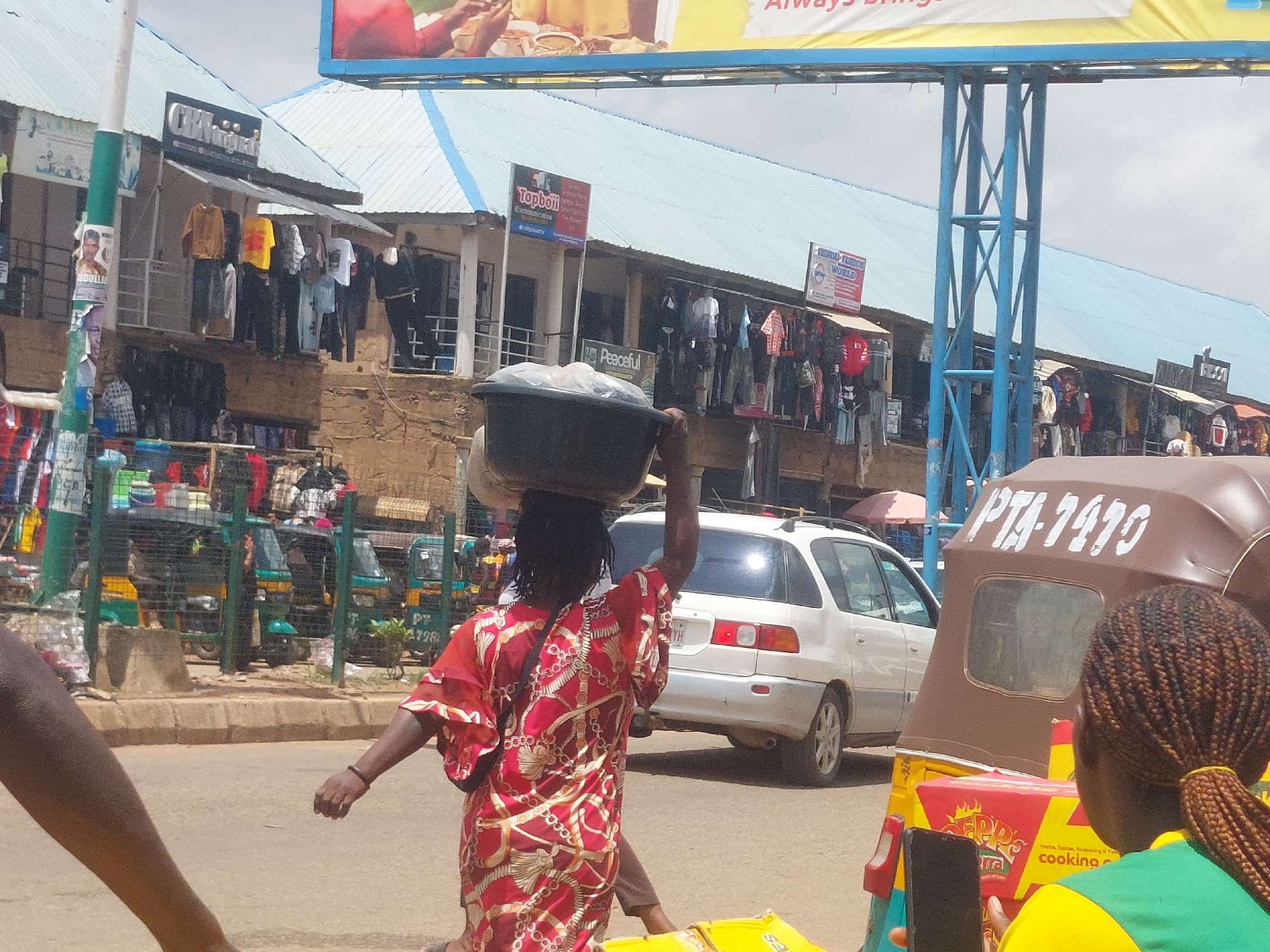 An outdoor, daytime scene depicts a street market in an urban environment. In the foreground, a person with dark skin and a red and gold patterned garment walks away from the camera, carrying a large dark bowl on their head. To the left, another person's arm and leg are visible, also in the foreground. In the middle ground, a white minivan is parked on the right side of the street, and a brown auto-rickshaw with the license plate "PTA-1415" is partially visible on the right. A green fence separates the market stalls from the street. The market stalls themselves display various clothing items, and signage is visible on some of the buildings. In the background, several other auto-rickshaws are parked. The sky is overcast.