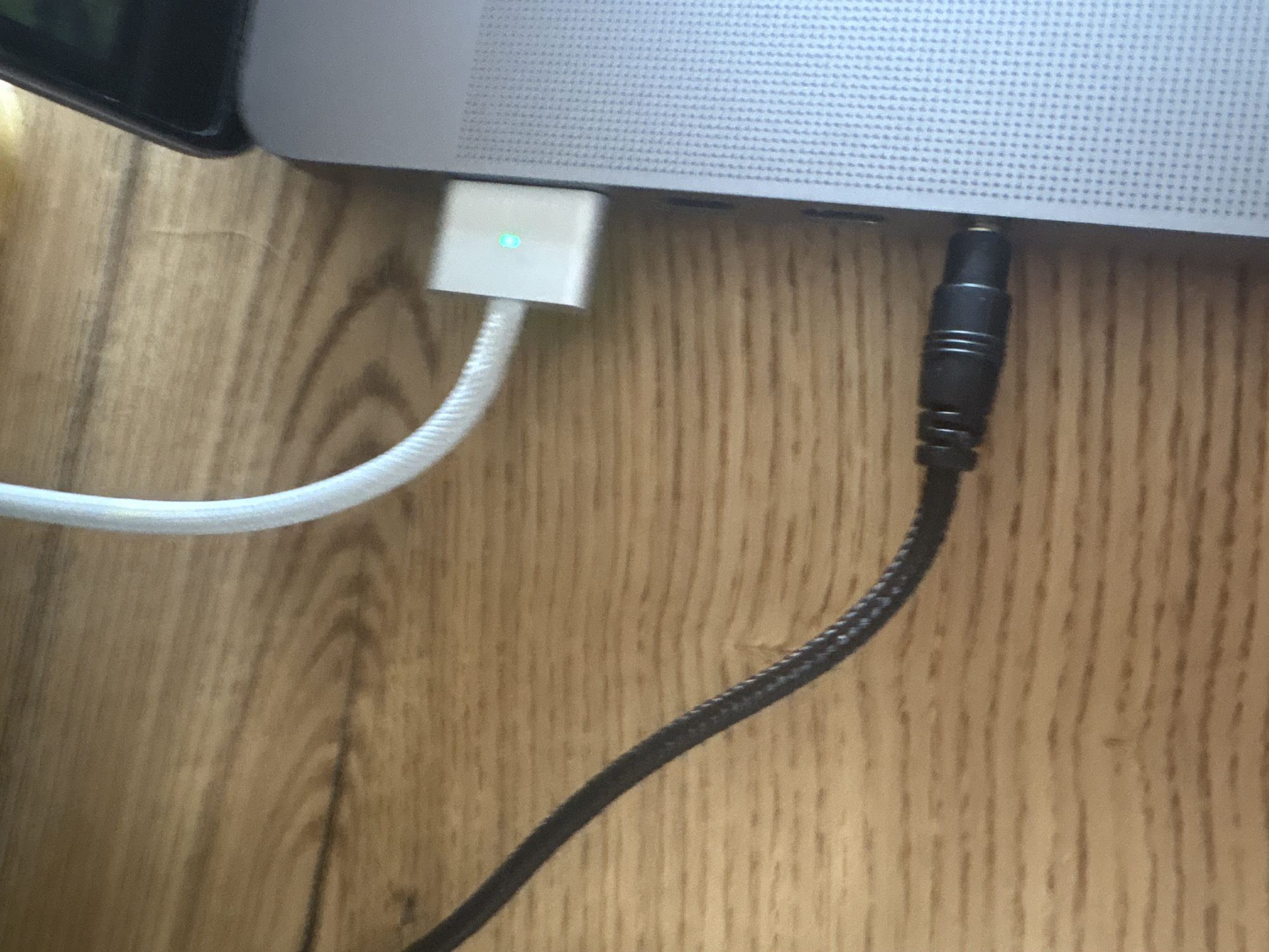 A close-up, eye-level shot shows a portion of a laptop and a wooden surface. On the left, a black audio cable is plugged into a port on the side of the laptop. The cable has a braided texture. To the right of the audio cable, a white USB-C cable is connected to a small white adapter, which is also plugged into the laptop. This adapter has a small, glowing green indicator light. The laptop itself is silver with a textured surface. The background is a wooden surface with visible grain patterns. The image appears to be taken indoors, likely on a desk or table. The location of Ореховка, Russia, is not depicted in the image.